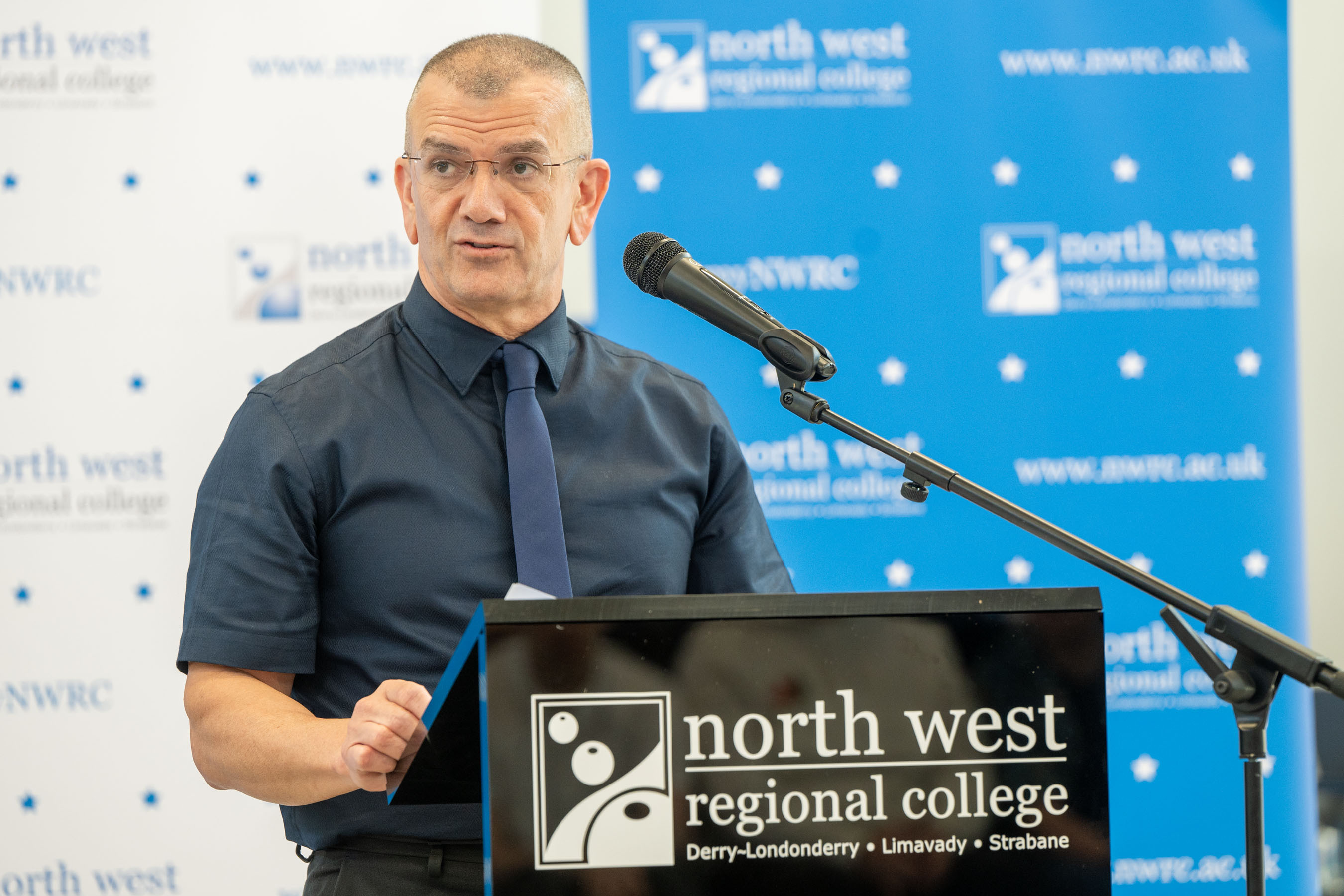 Speaker in front of podium at college celebration event. There is college branding for NWRC in the background.