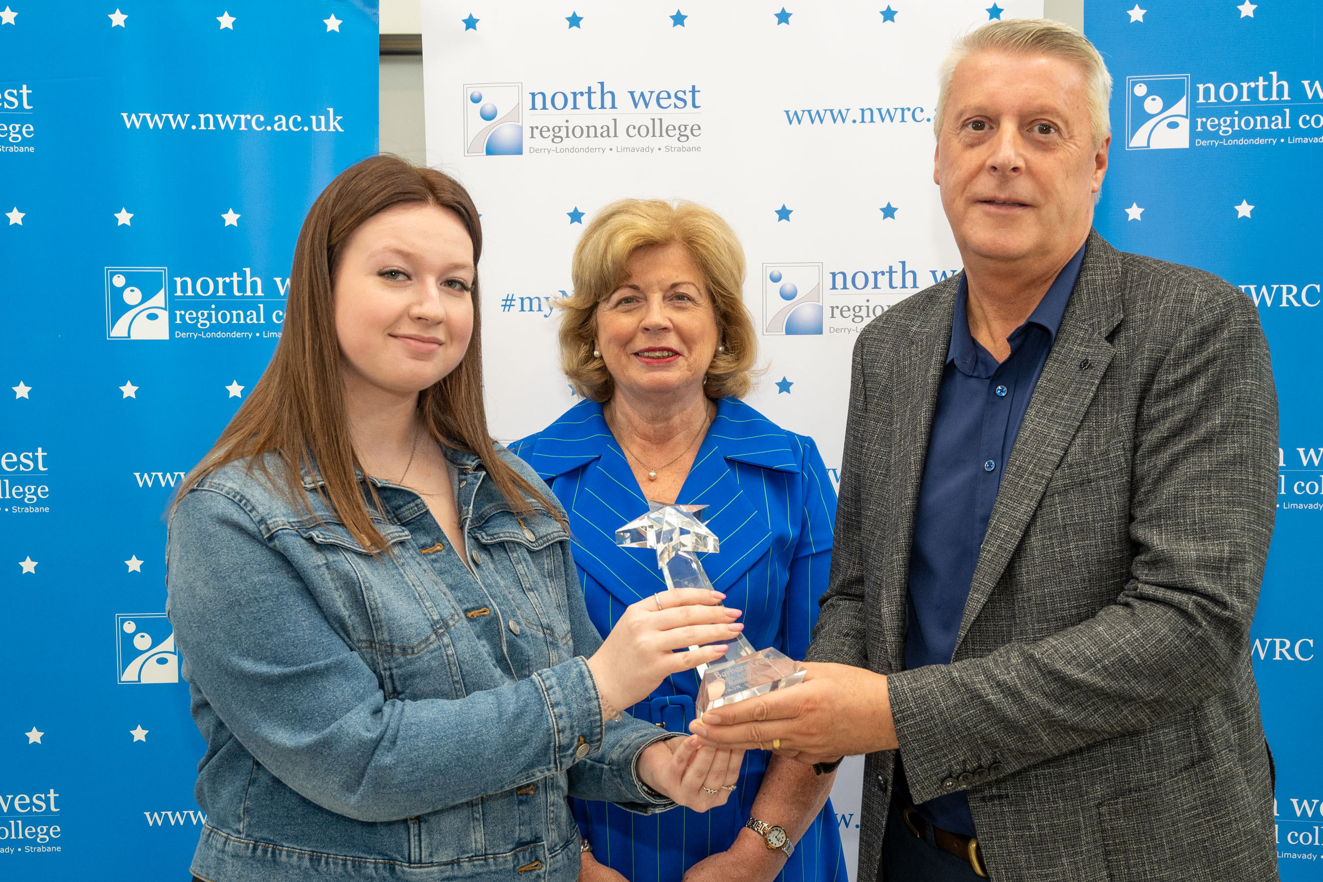 Two women and a man in front of NWRC pop ups. The man is presenting one of the women with a trophy.