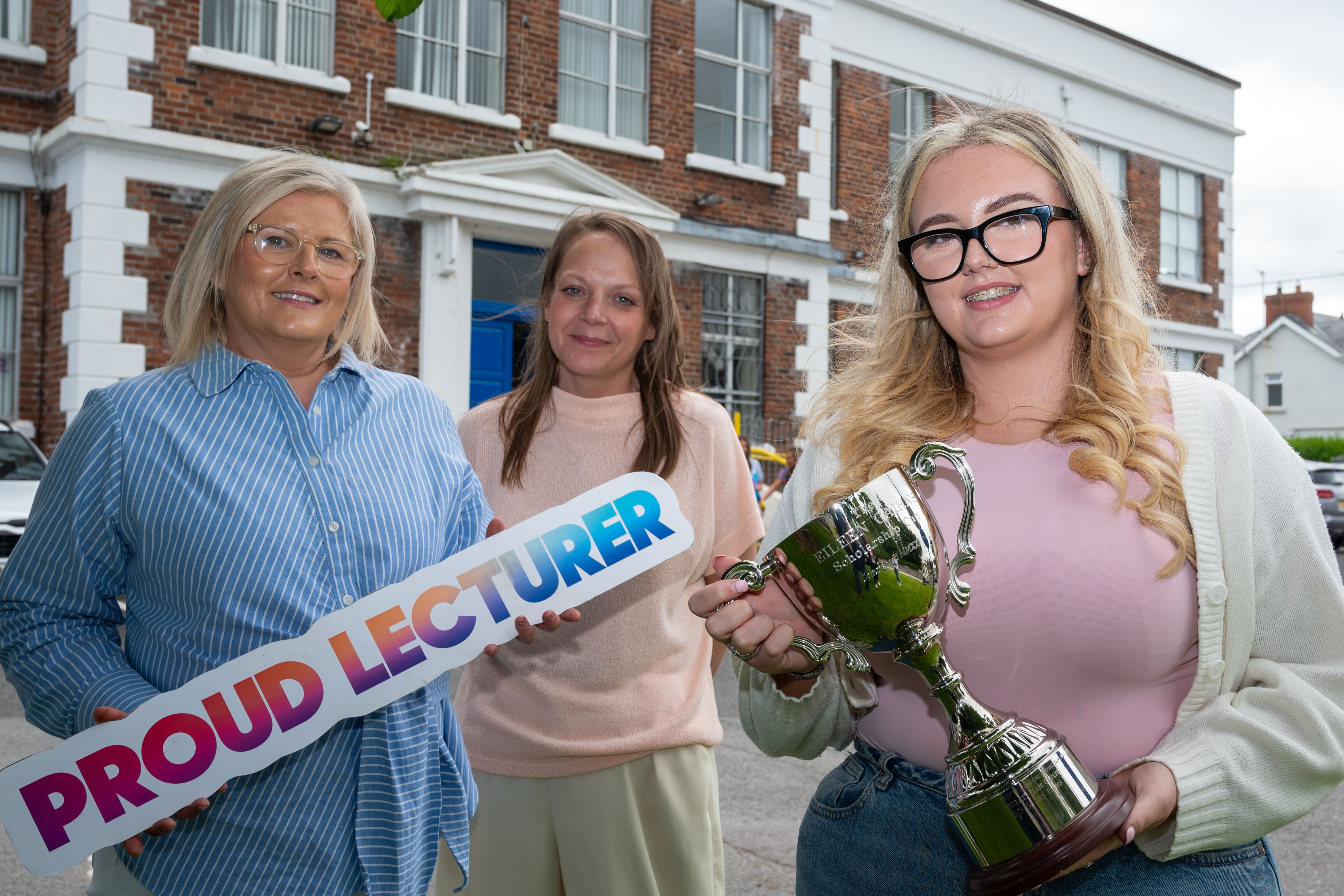 Three people at celebration event. One is holding a trophy, one has a proud lecturer sign.