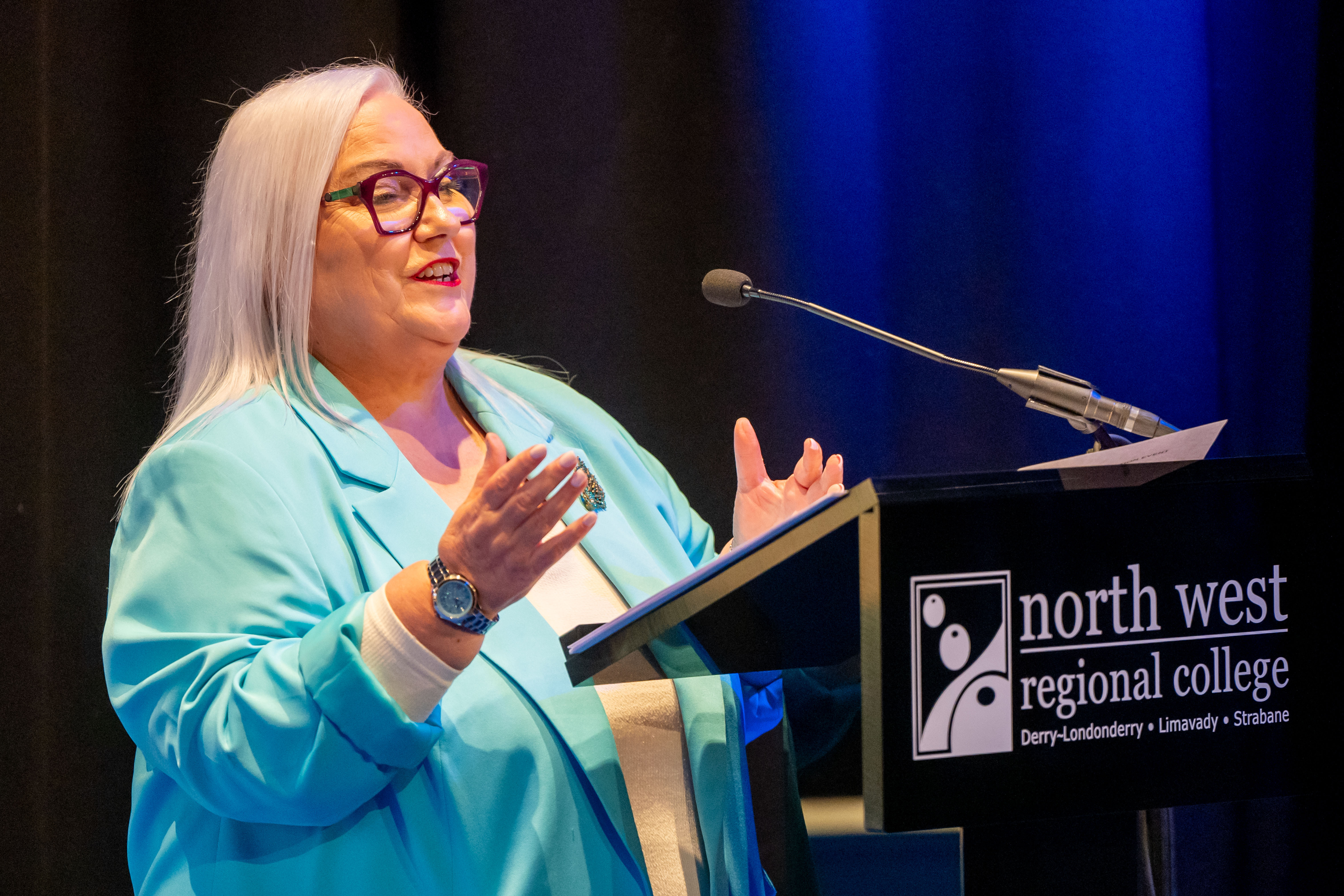 Speaker in front of podium at college celebration event. There is college branding for NWRC in the background.