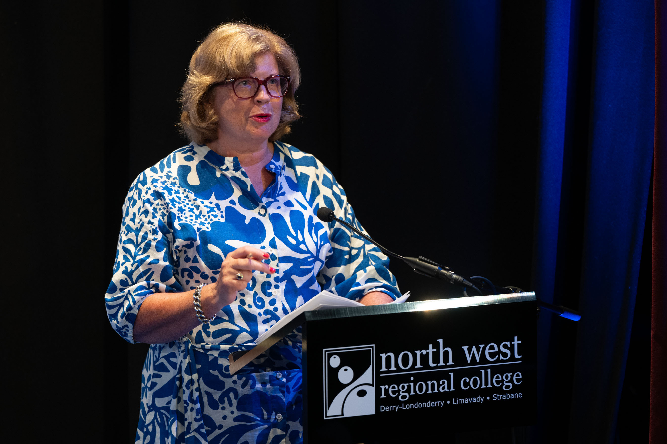 Speaker in front of podium at college celebration event. There is college branding for NWRC in the background.