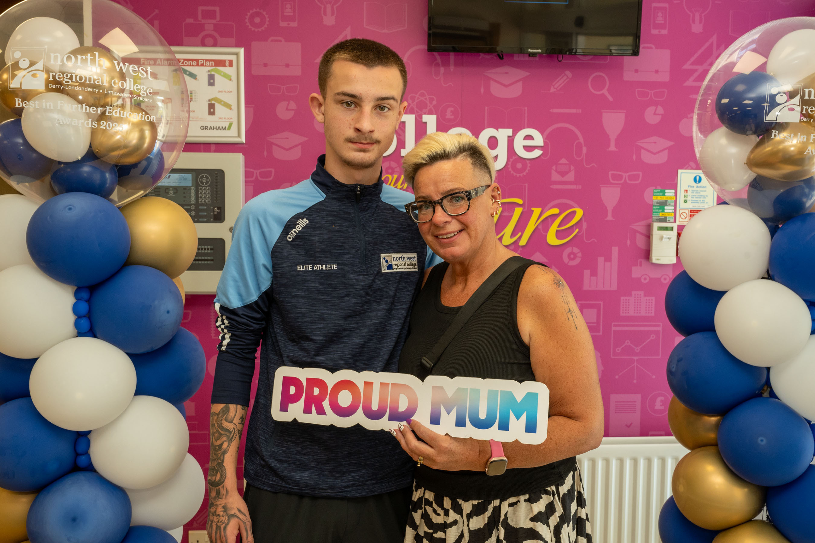 Two people at celebration event. One is holding a certificate, one has a proud mum sign.