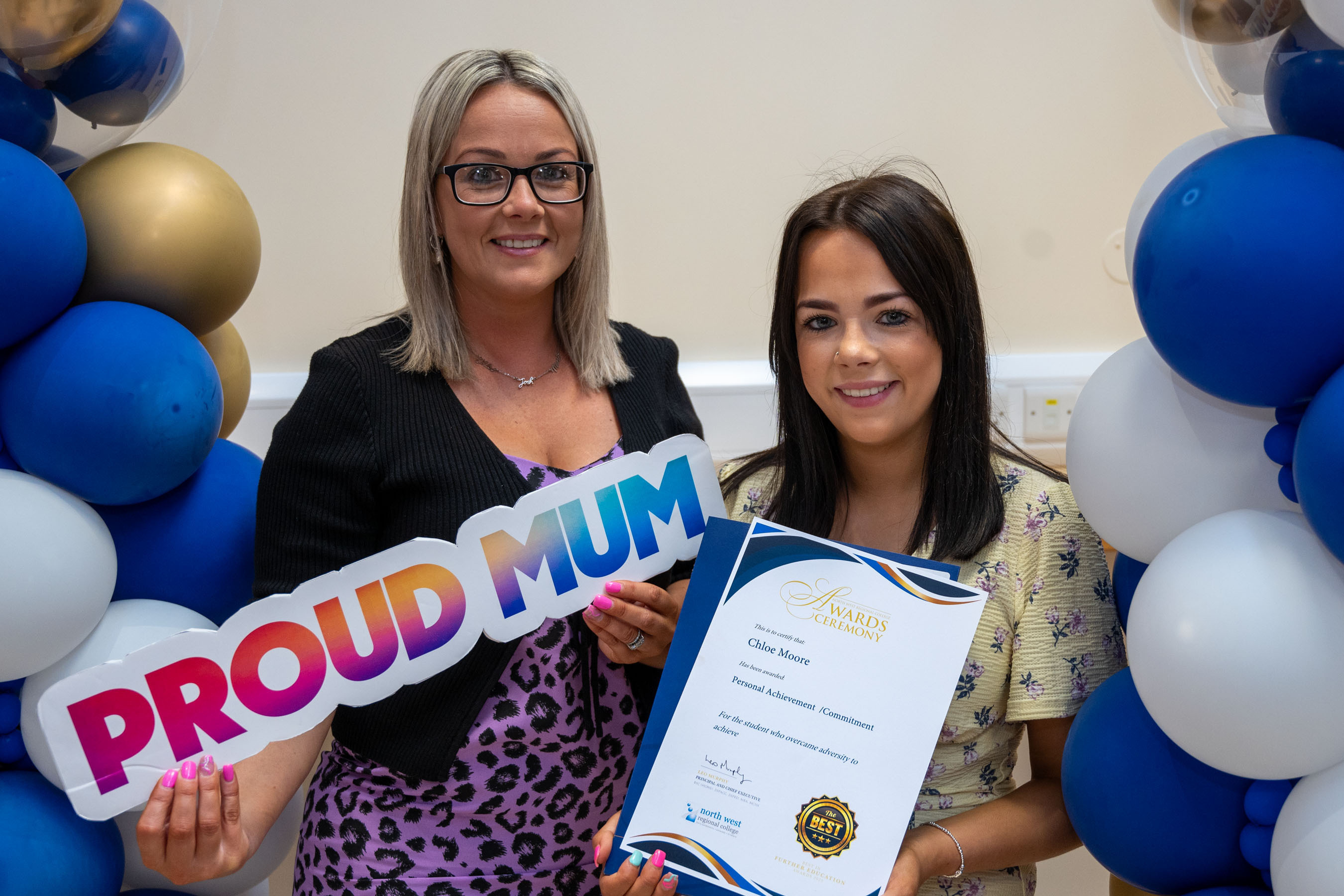Two people at celebration event. One is holding a certificate, one has a proud mum sign.