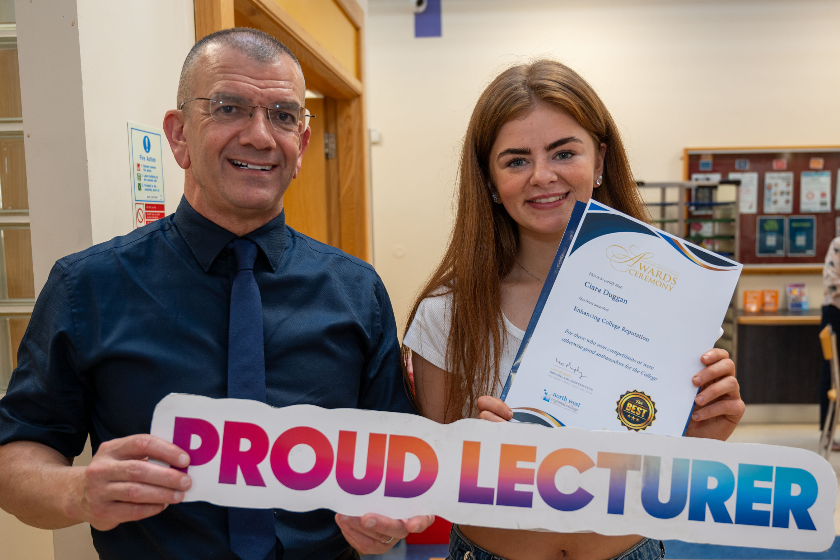 Two people at celebration event. One is holding a certificate, one has a proud lecturer sign.