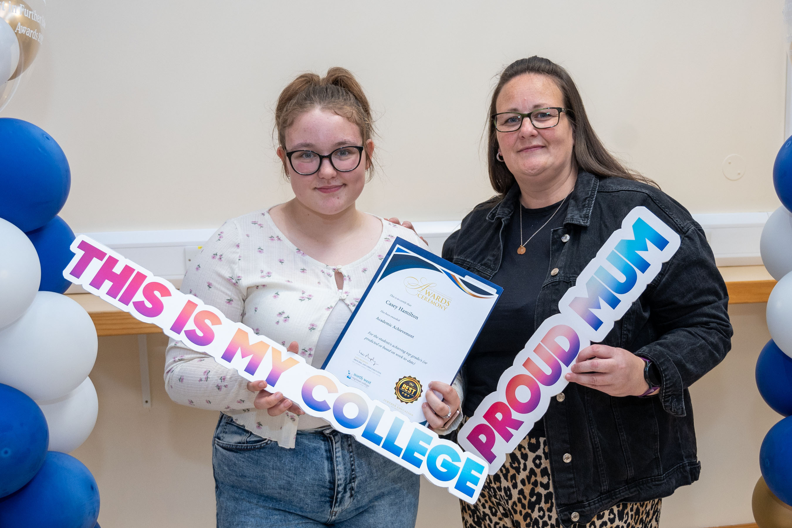 Two people at celebration event. One is holding a certificate, one has a proud mum sign.
