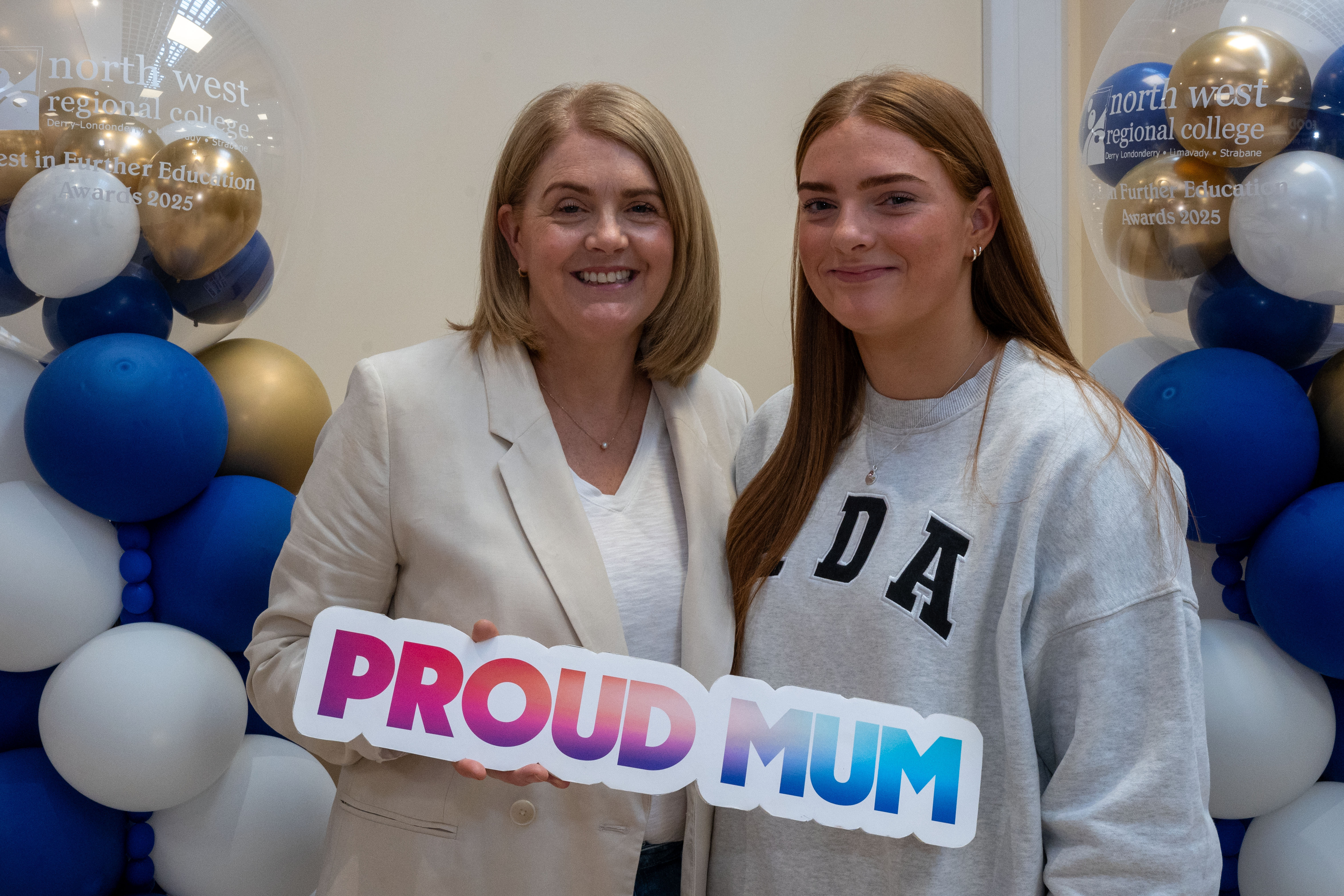 Two people at celebration event. One is holding a certificate, one has a proud mum sign.