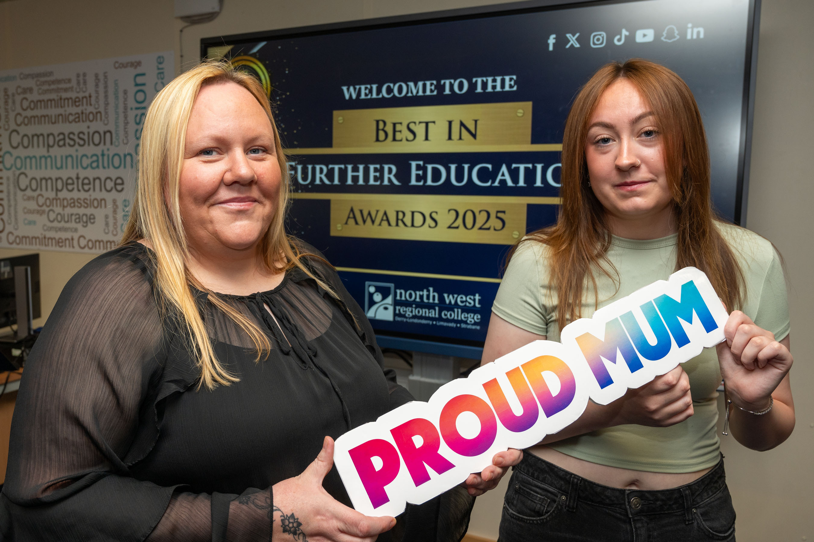 Two people at celebration event. One is holding a sign that says proud mum