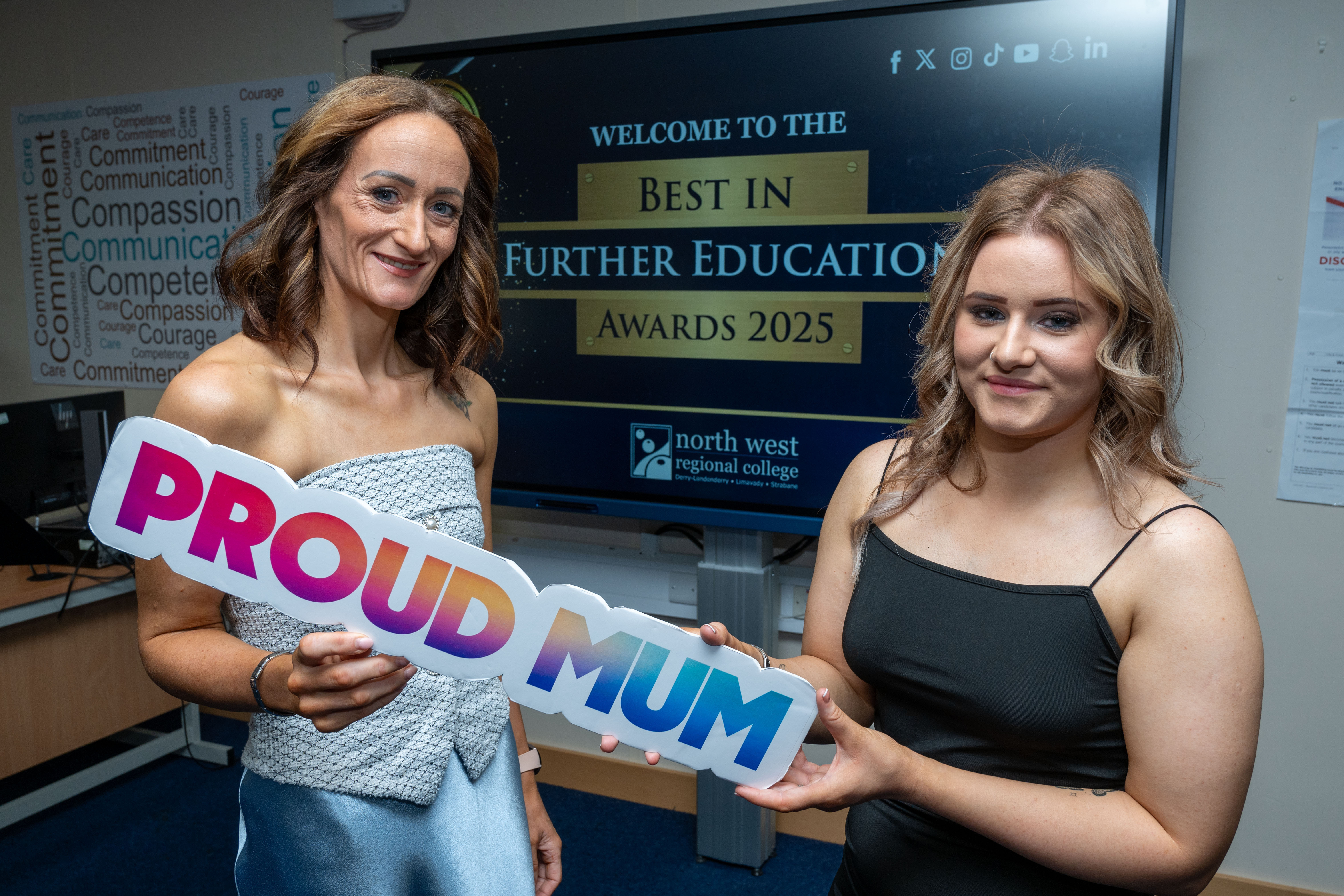 Two people at celebration event. One is holding a sign that says proud mum