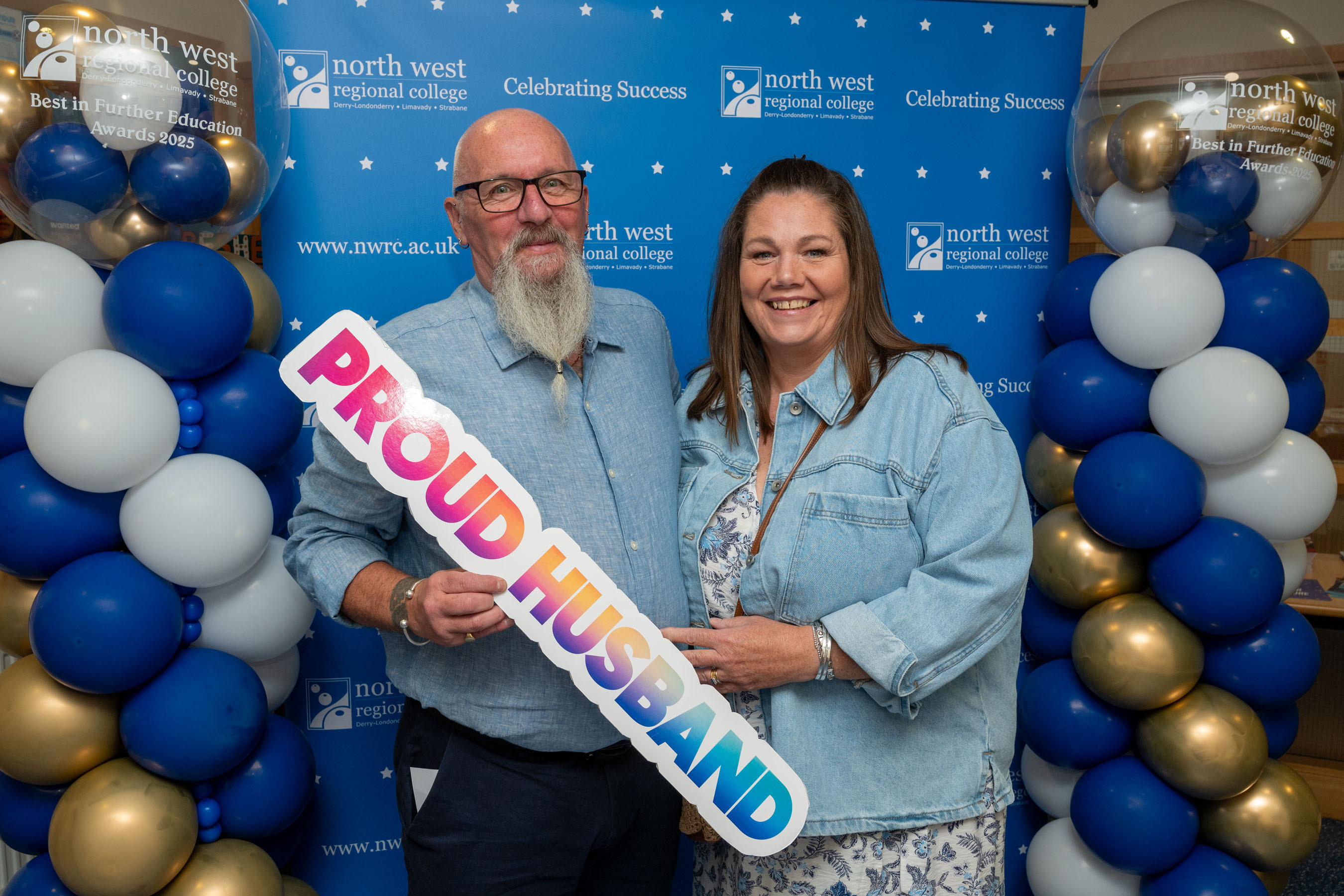 Two people at celebration event. One is holding a sign that says proud husband