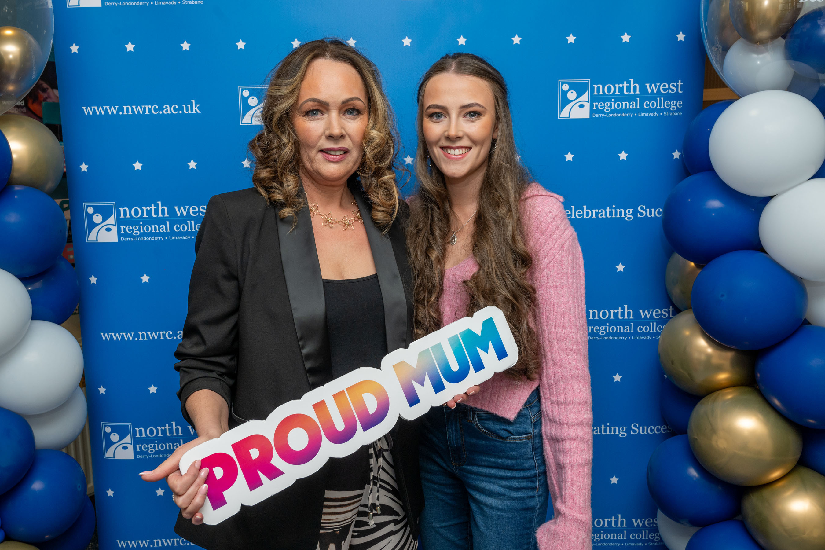 Two people at celebration event. One is holding a sign that says proud mum