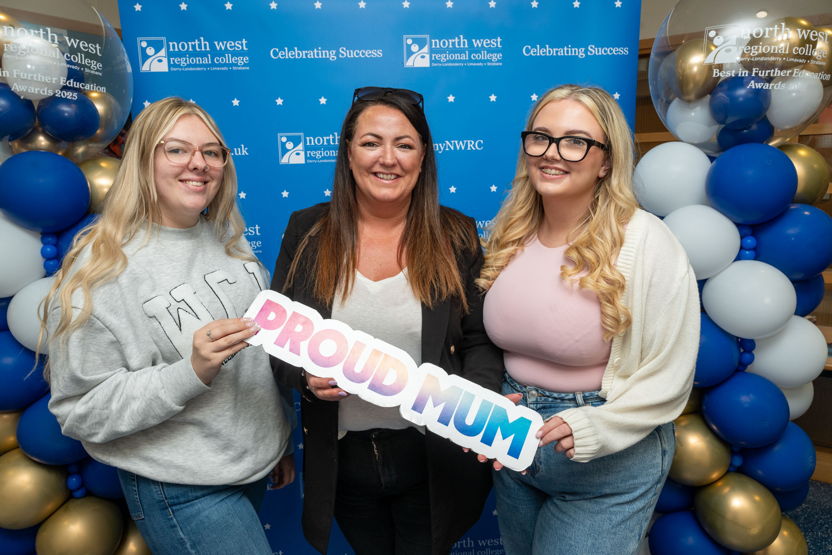 Three people at celebration event. One is holding a sign that says proud mum