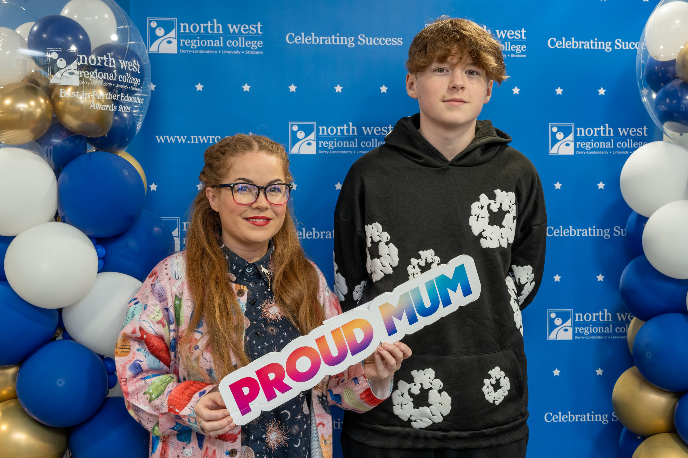 Two people at celebration event. One is holding a sign that says proud mum
