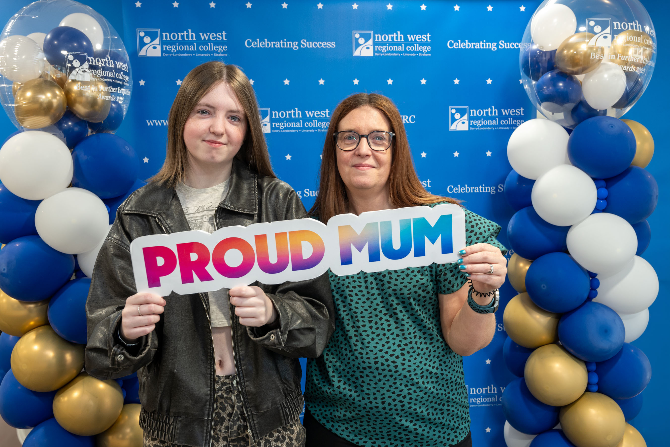 Two people at celebration event. One is holding a sign that says proud mum
