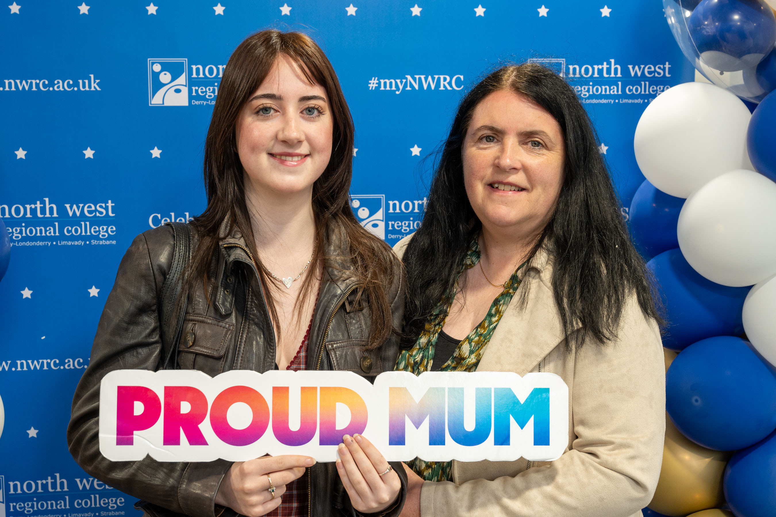 Two people at celebration event. One is holding a sign that says proud mum