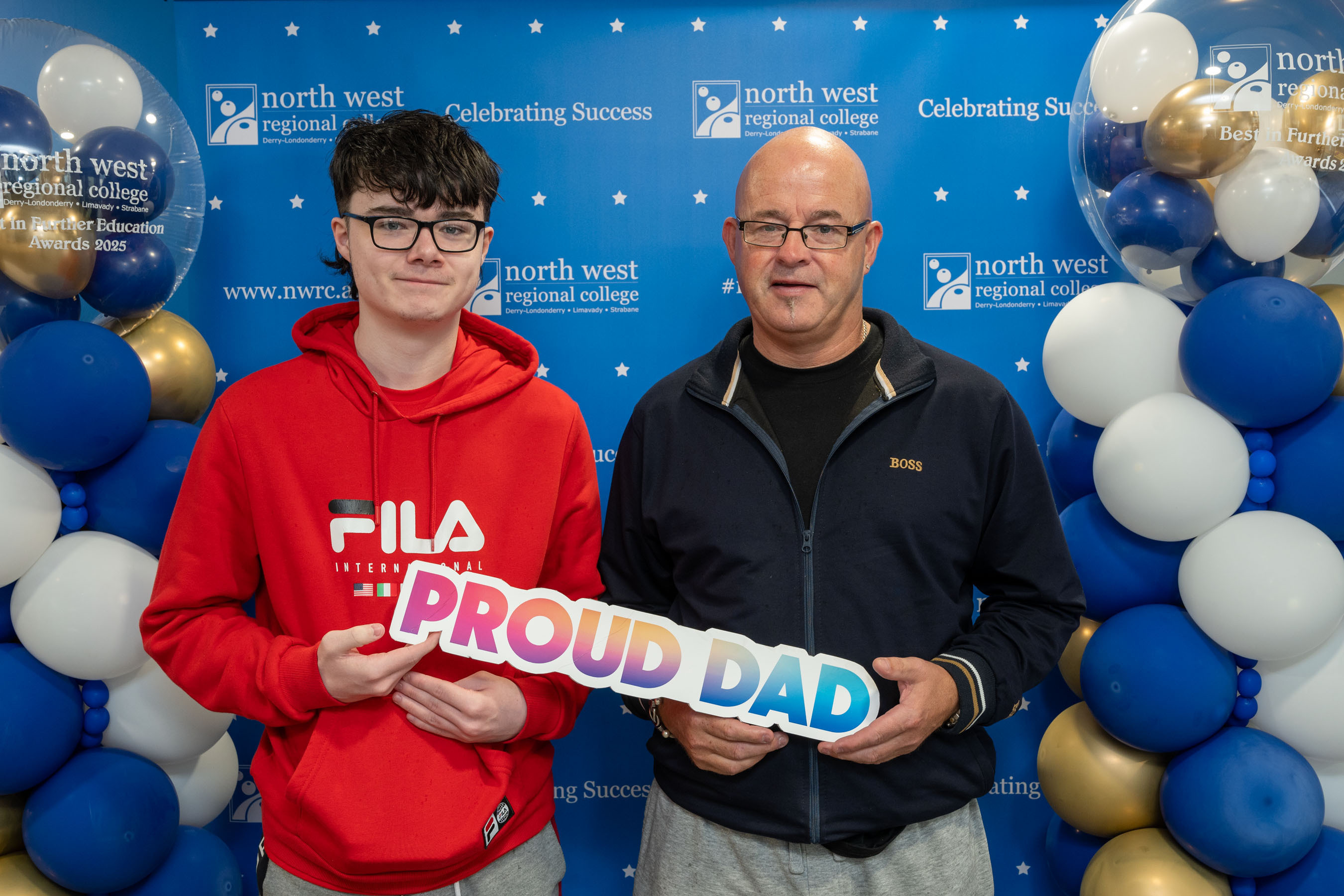 Two people at celebration event. One is holding a sign that says proud dad