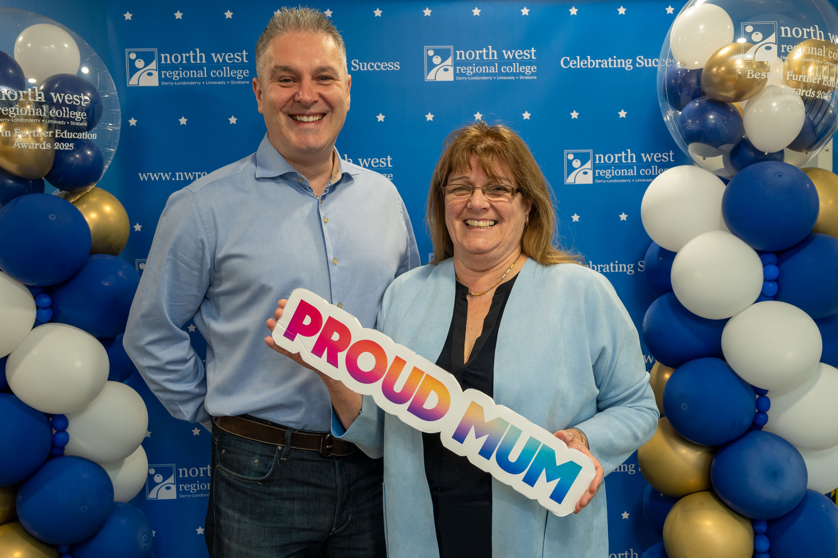Two people at celebration event. One is holding a sign that says proud mum
