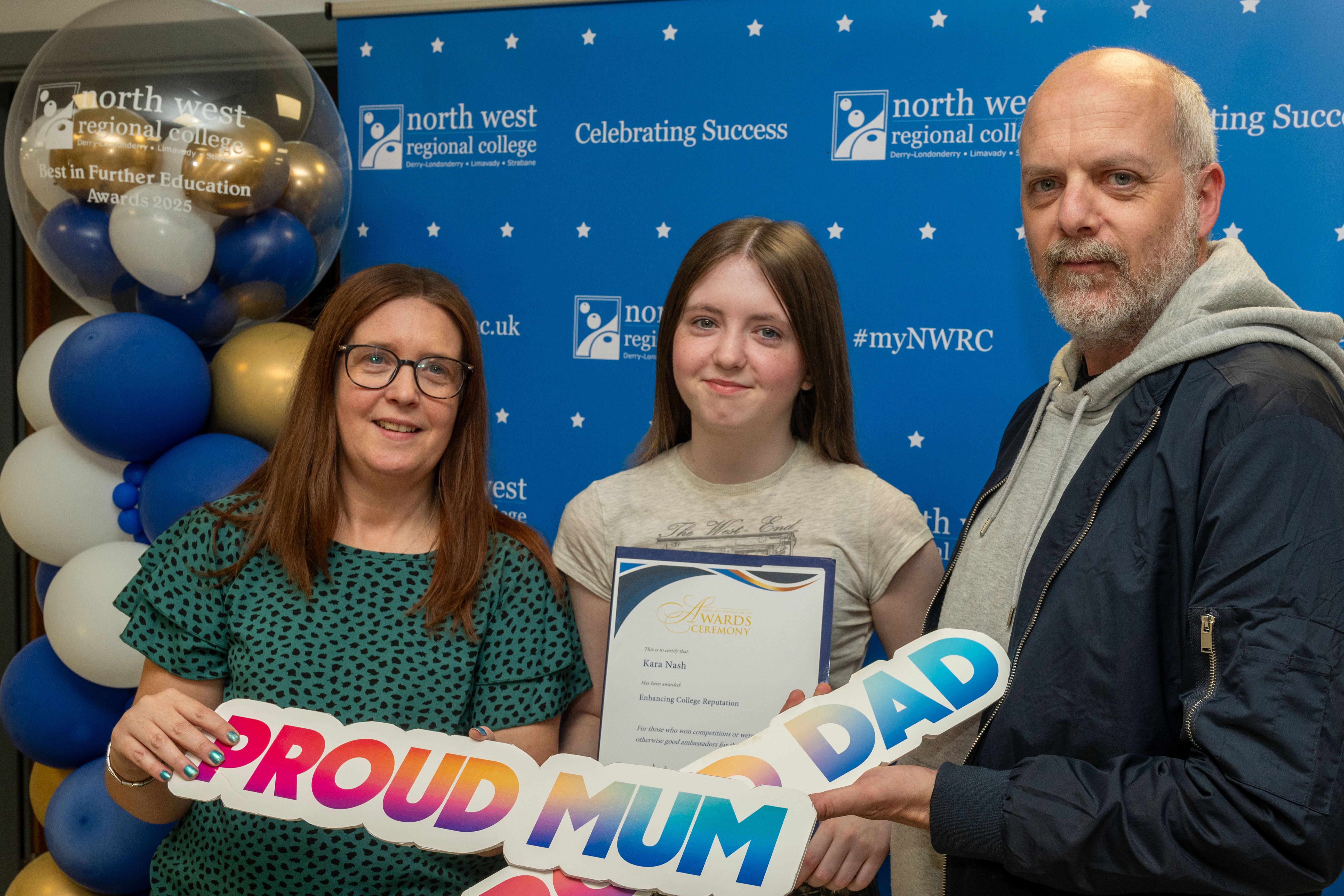 Three people at celebration event. One is holding a sign that says proud mum and the other with a sign for proud dad