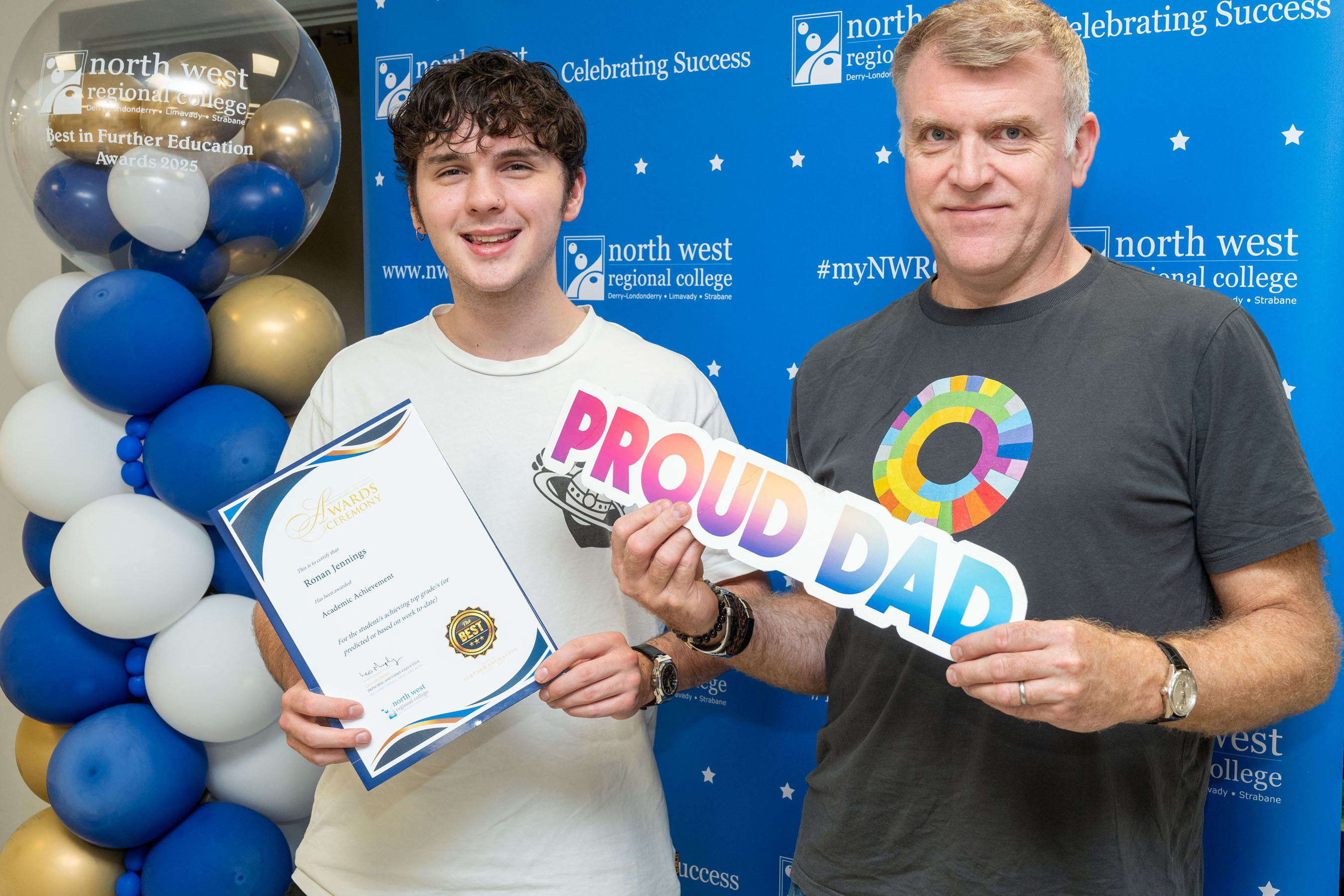 Two people at celebration event. One is holding a sign that says proud dad