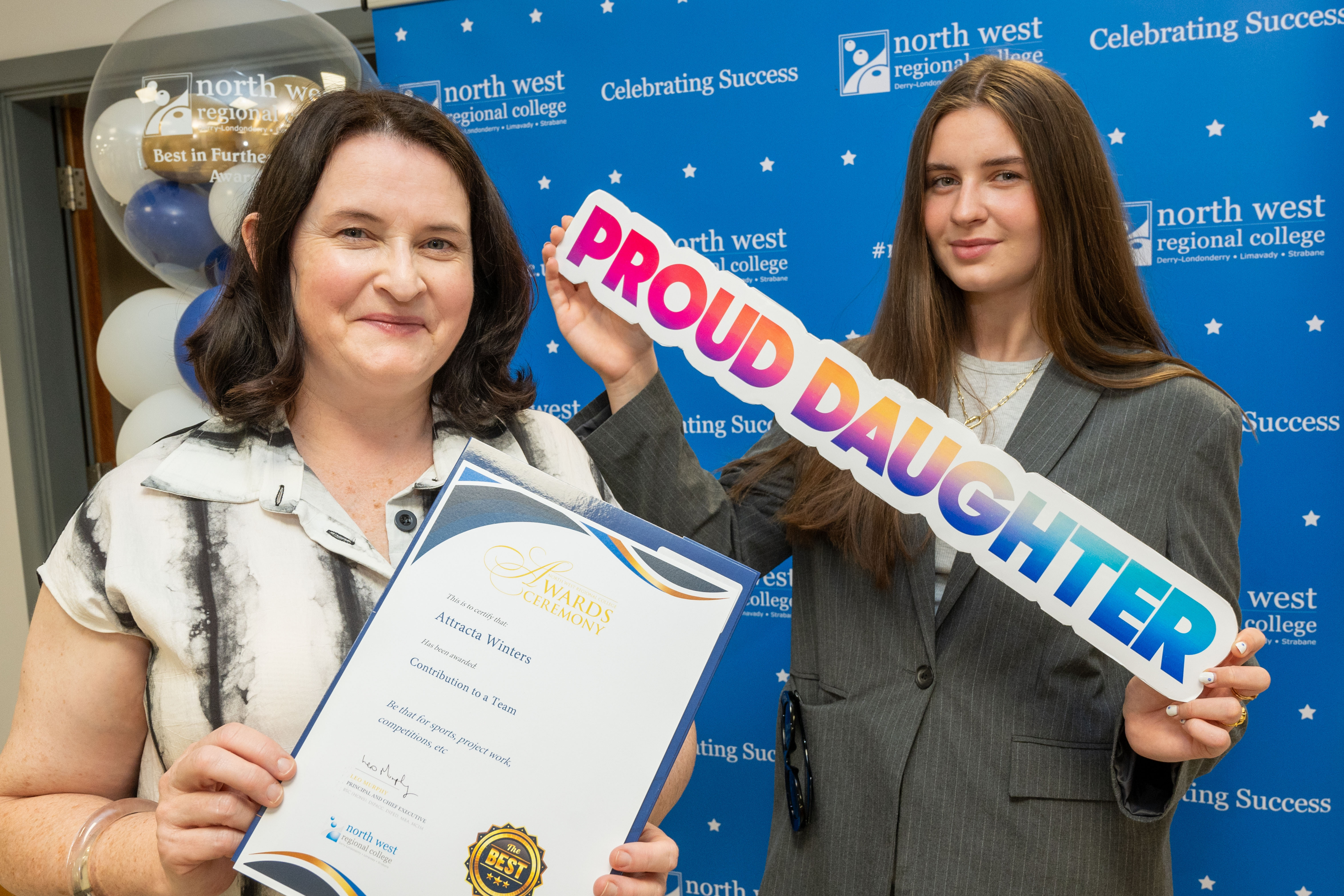 Two people at celebration event. One is holding a sign that says proud daughter