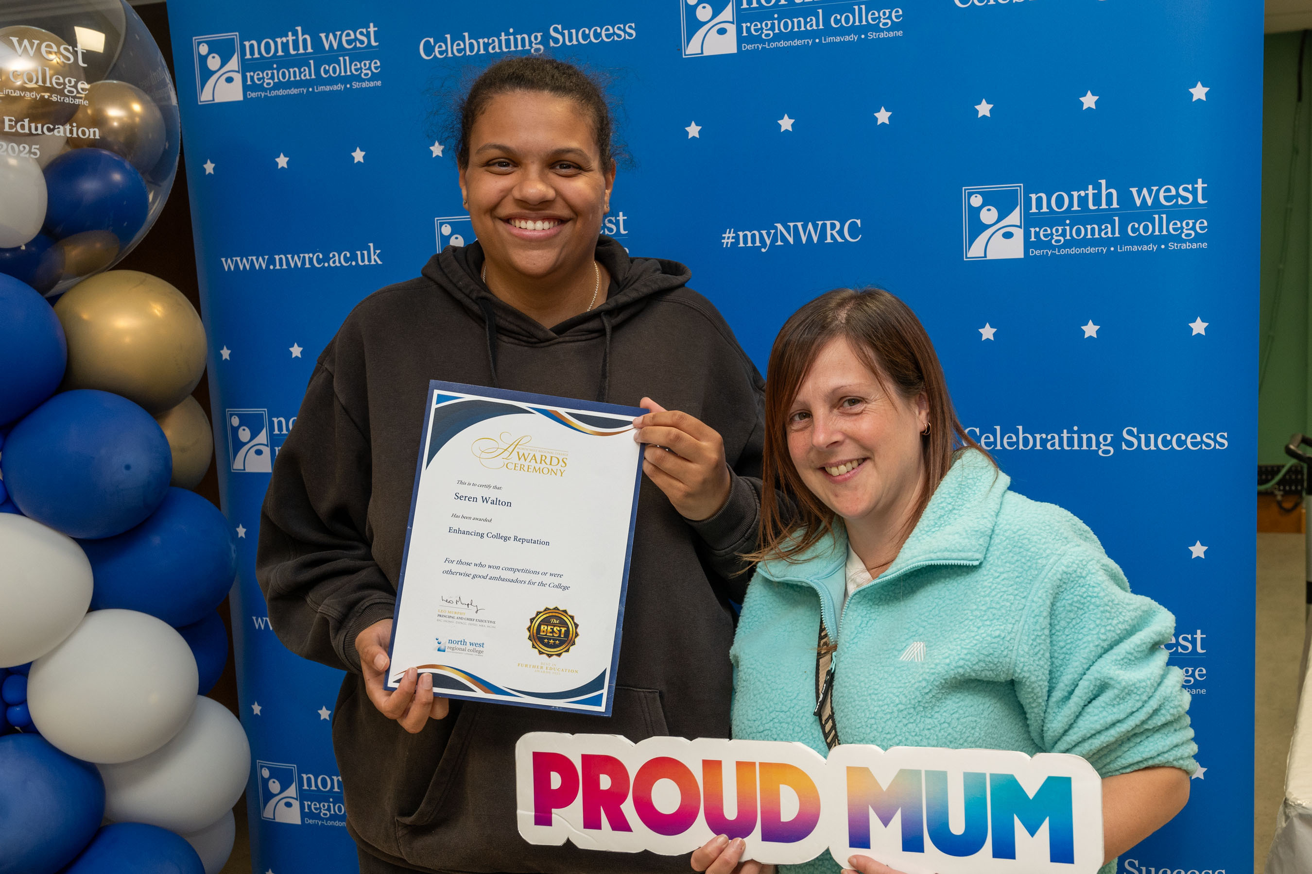Two people at celebration event. One is holding a sign that says proud mum