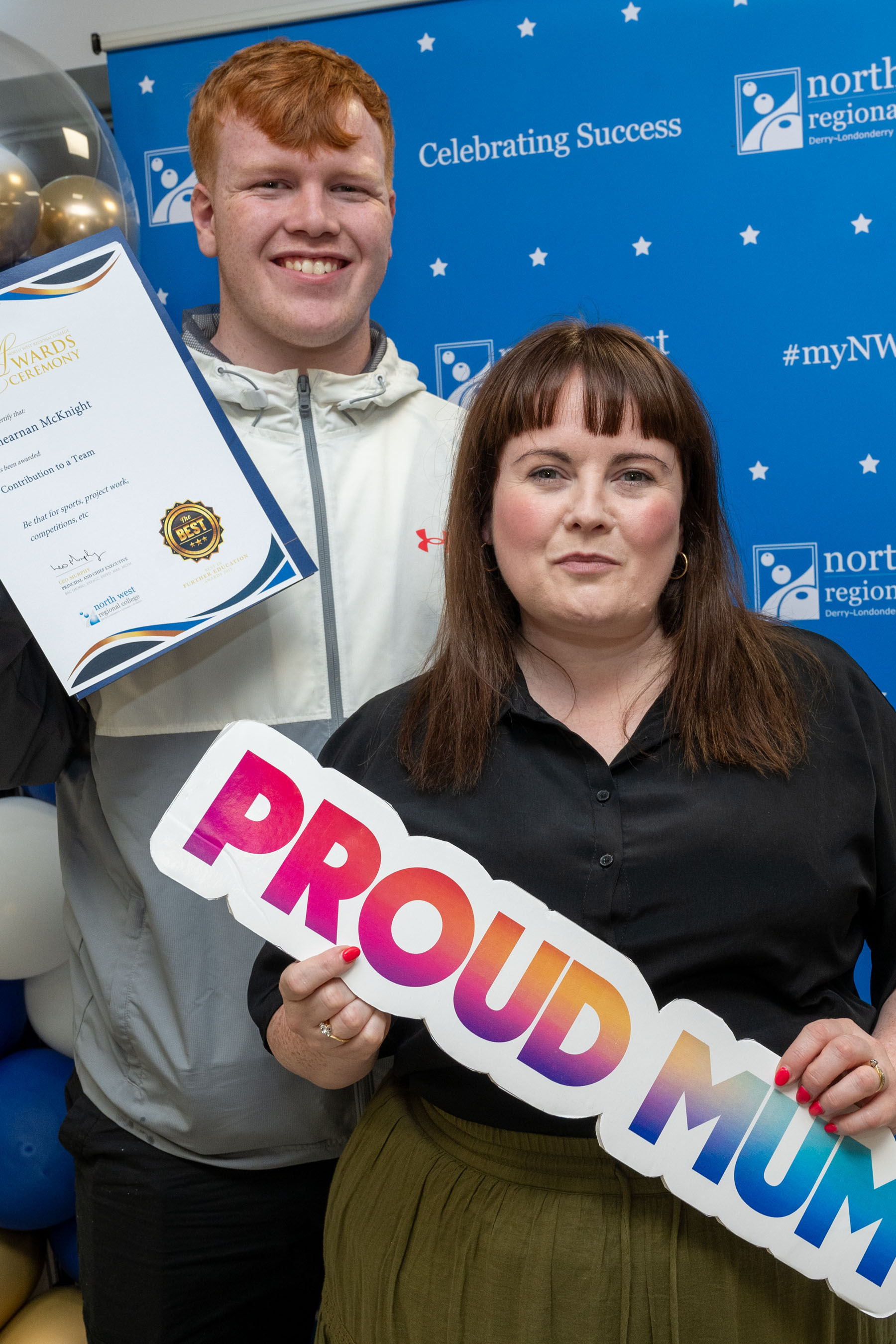 Two people at celebration event. One is holding a sign that says proud mum