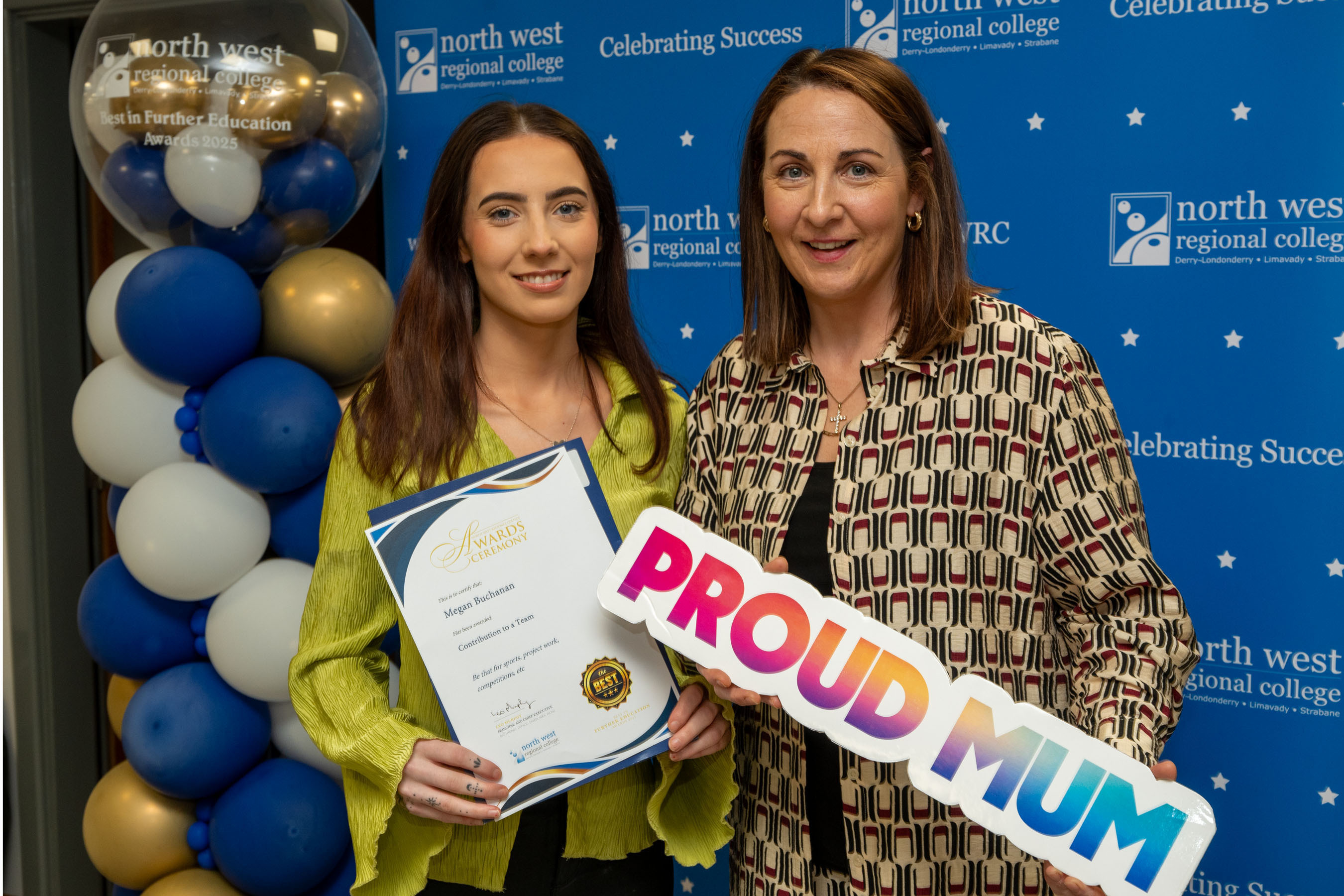 Two people at celebration event. One is holding a sign that says proud mum