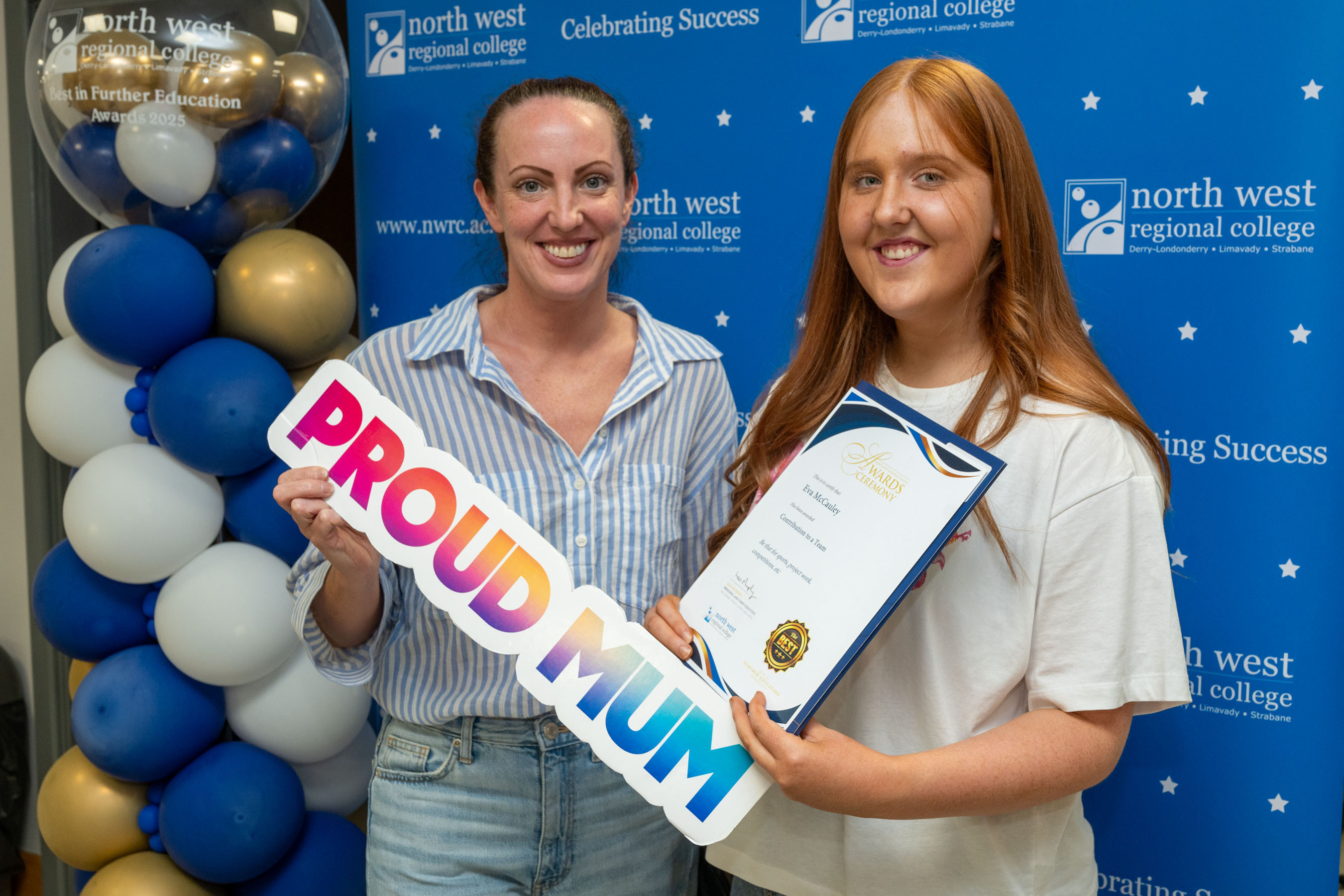 Two people at celebration event. One is holding a sign that says proud mum