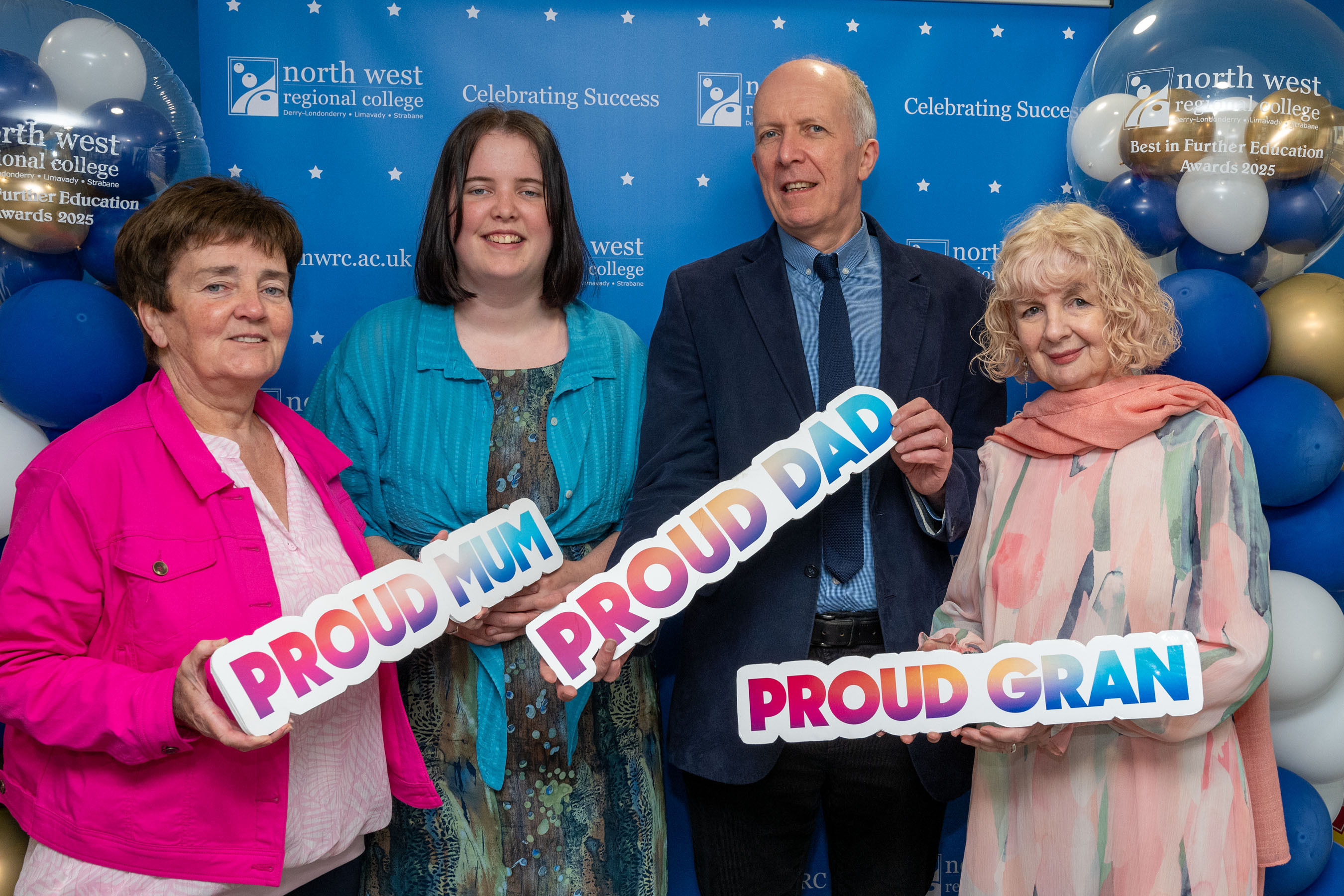 Four people at celebration event. One has a proud mum sign, one has a proud dad sign, one has a proud gran sign