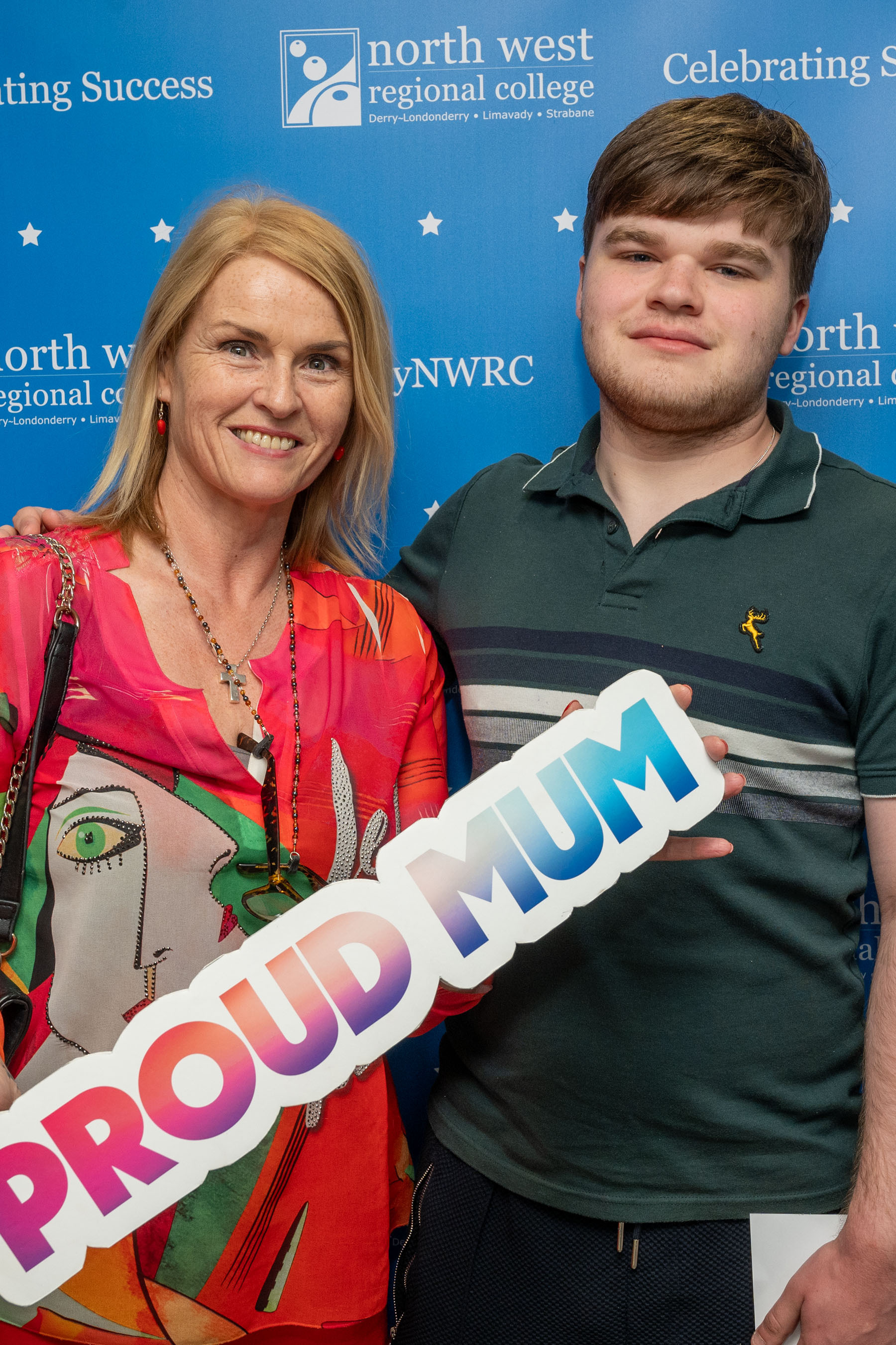 Two people at celebration event. One is holding a sign that says proud mum