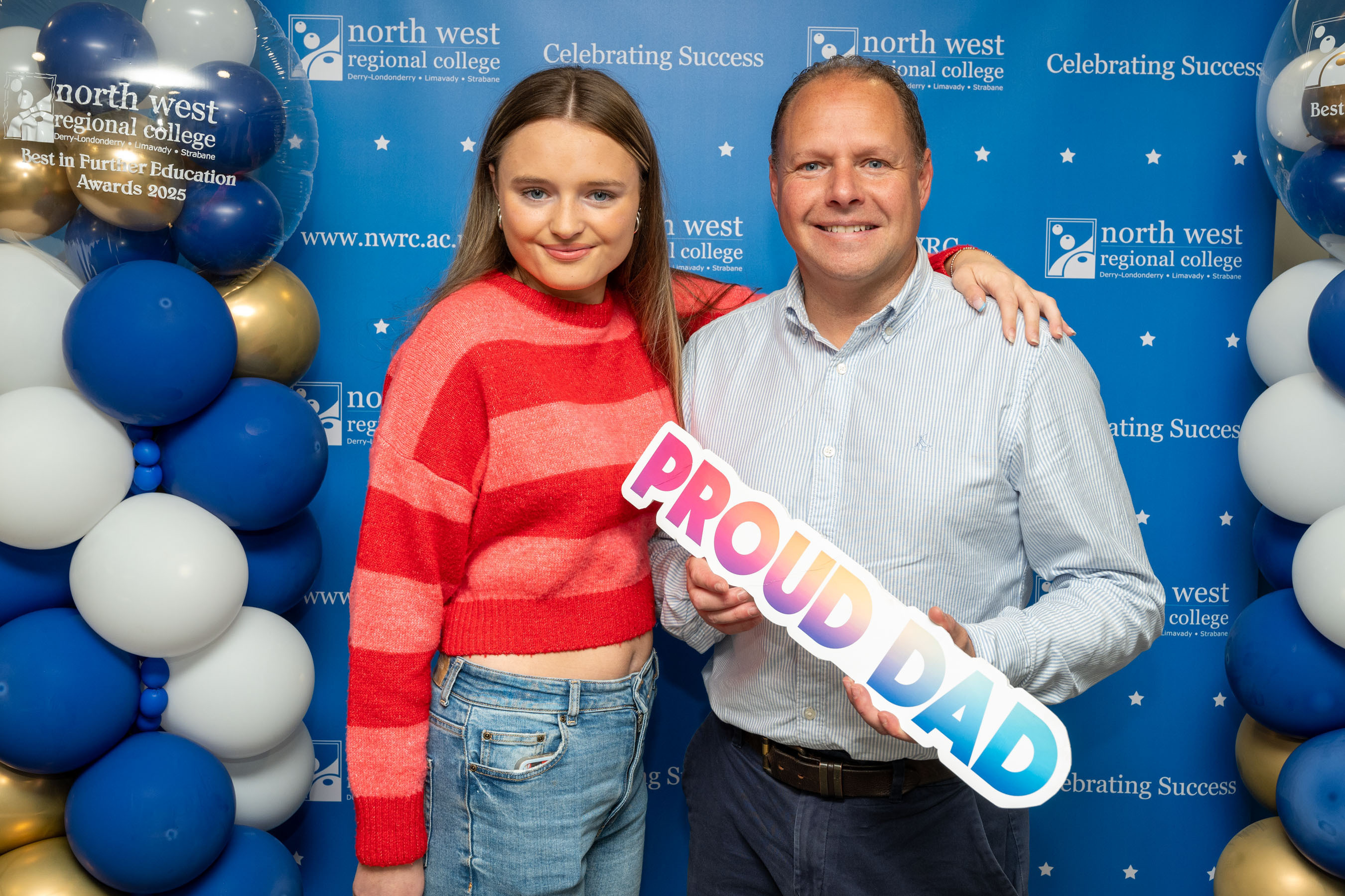 Two people at celebration event. One is holding a sign that says proud dad