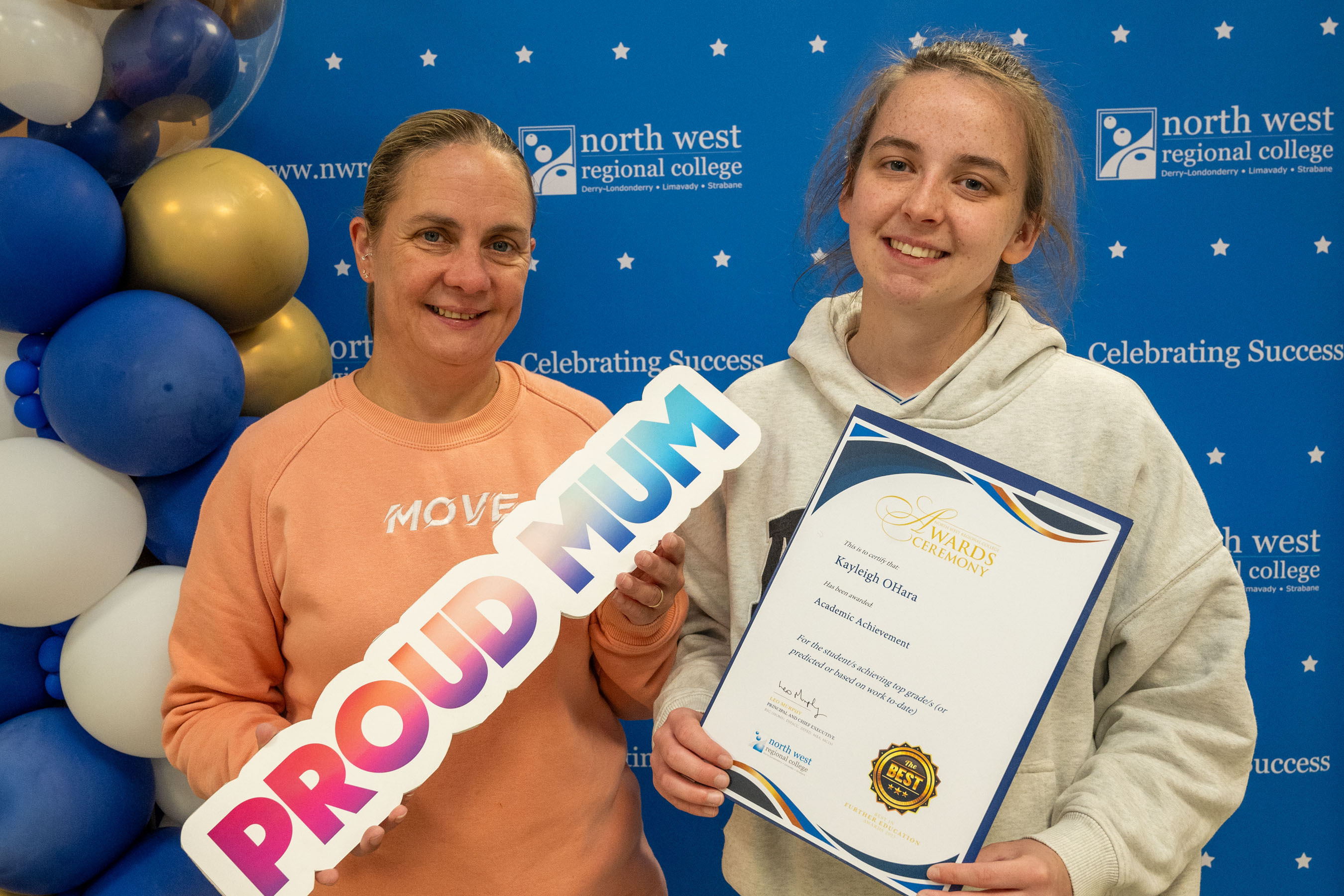 Two people at celebration event. One is holding a sign that says proud mum