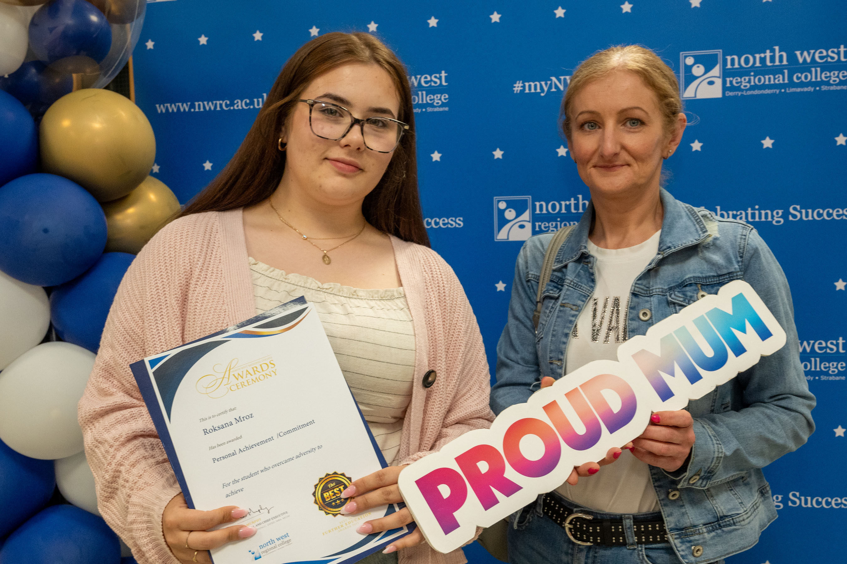 Two people at celebration event. One is holding a sign that says proud mum