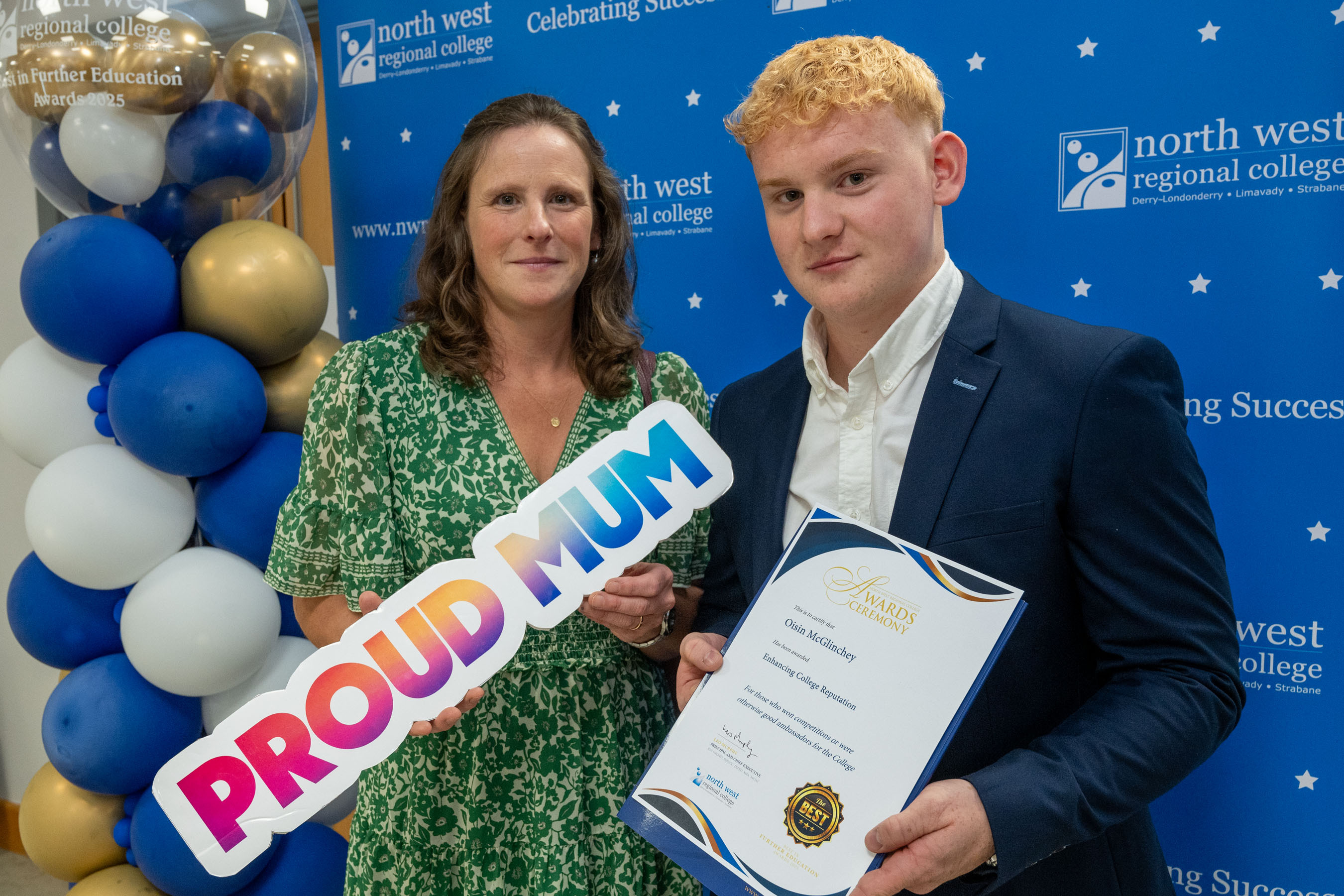Two people at celebration event. One is holding a sign that says proud mum