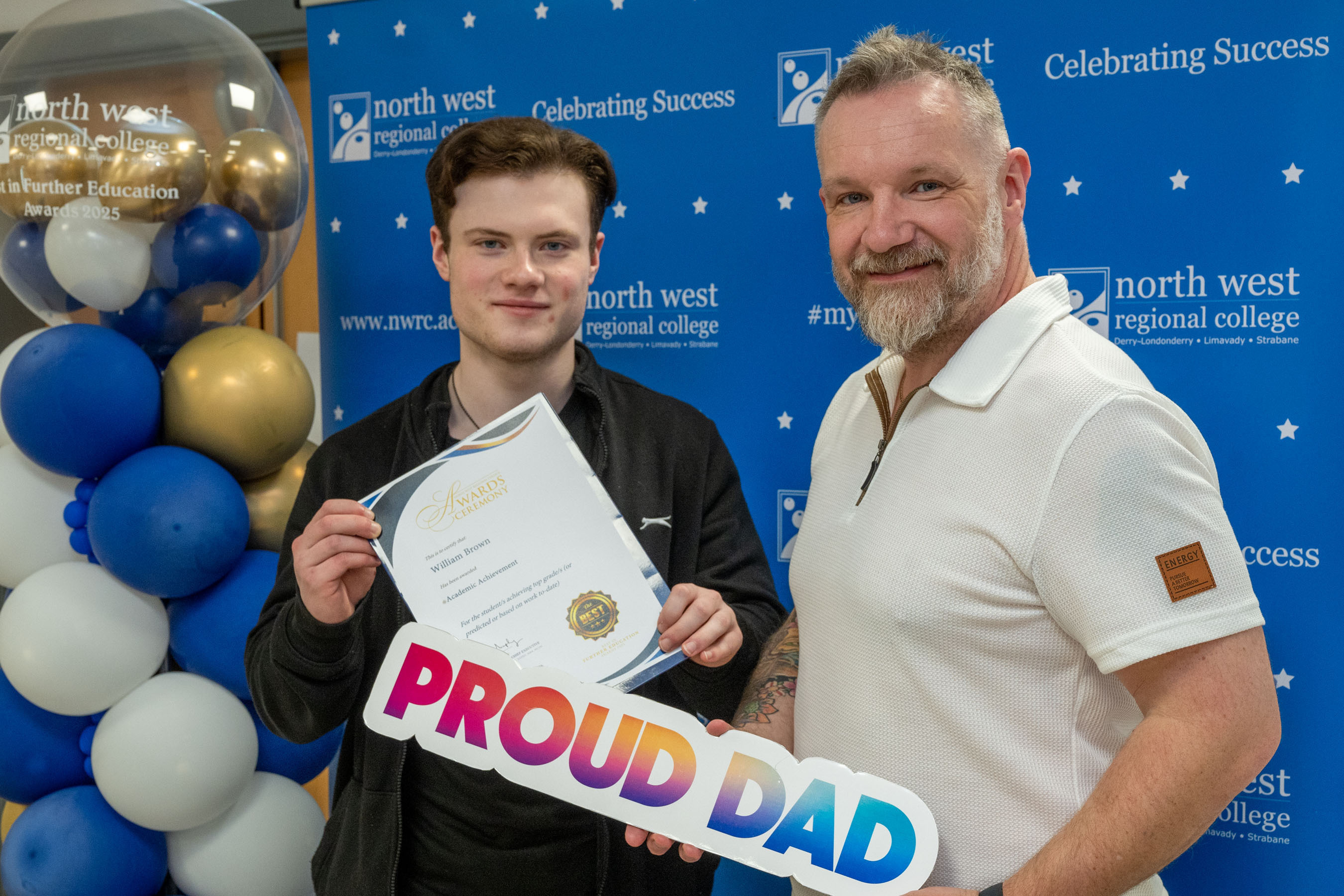 Two people at celebration event. One is holding a sign that says proud dad