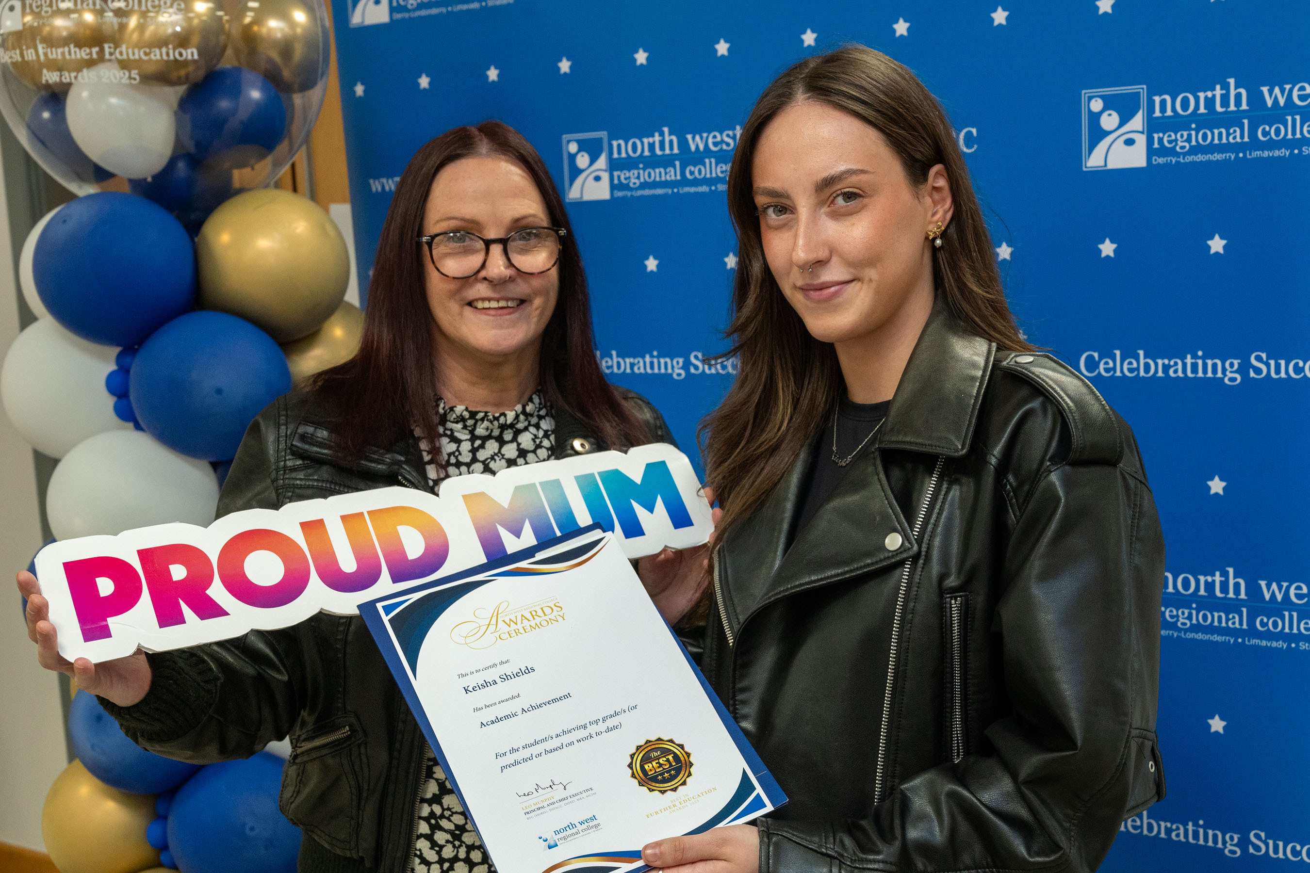 Two people at celebration event. One is holding a sign that says proud mum