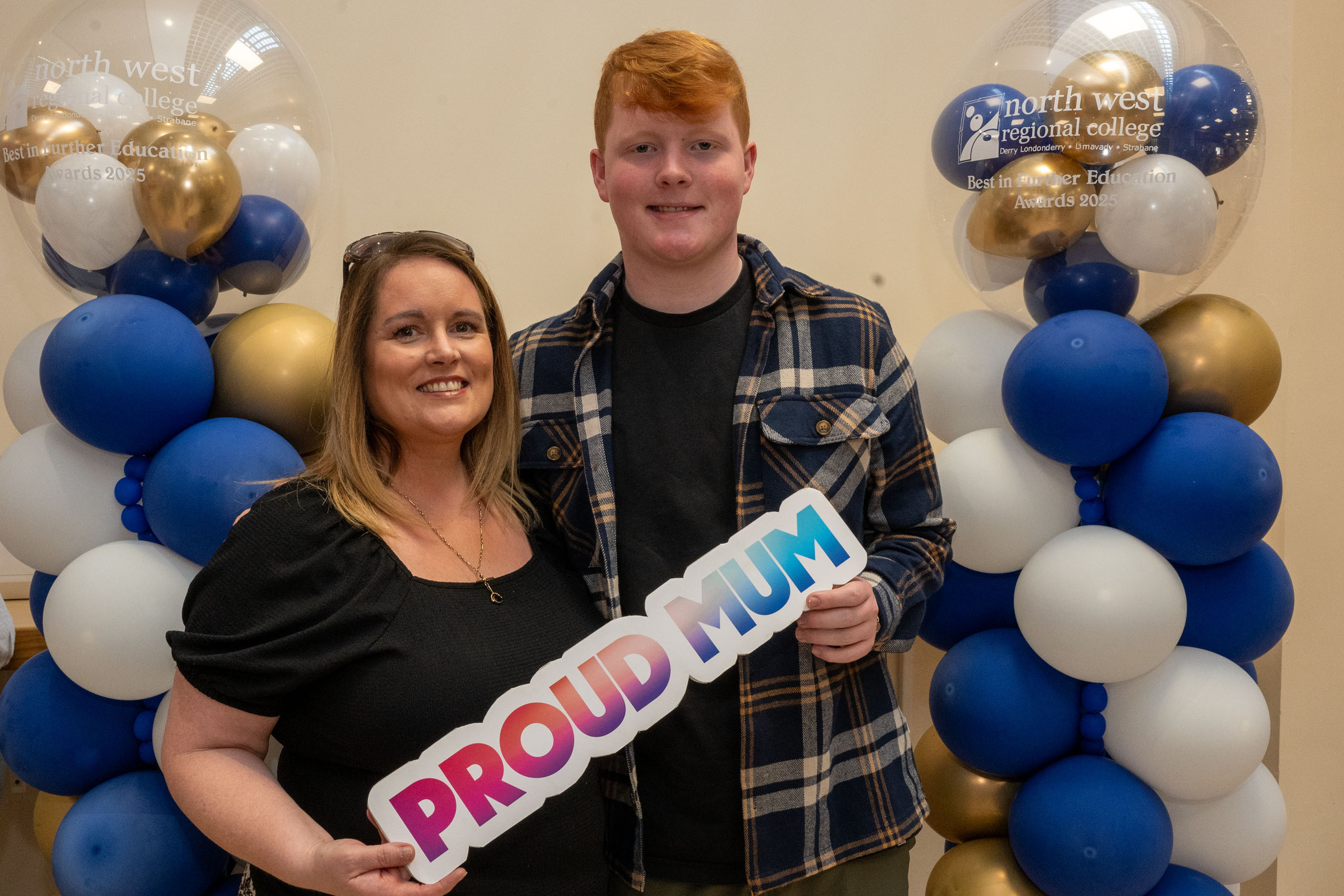 Two people at celebration event. One is holding a certificate, one has a proud mum sign.