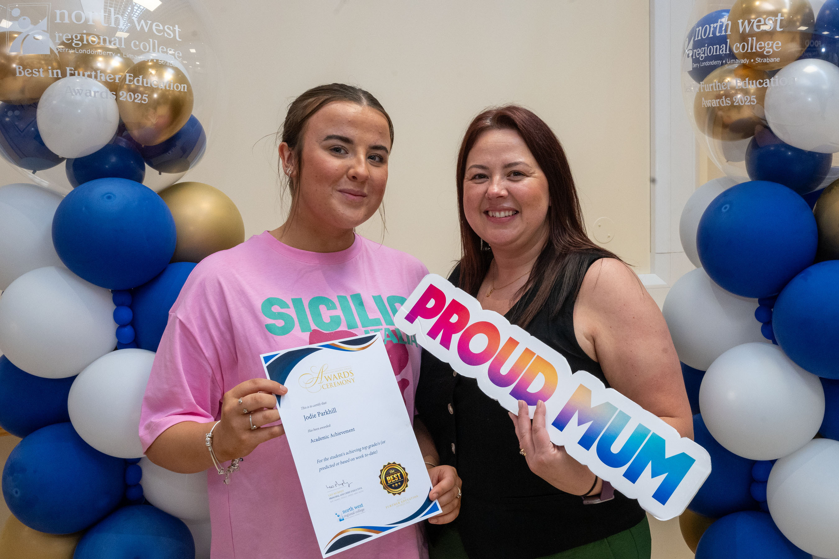 Two people at celebration event. One is holding a certificate, one has a proud mum sign.
