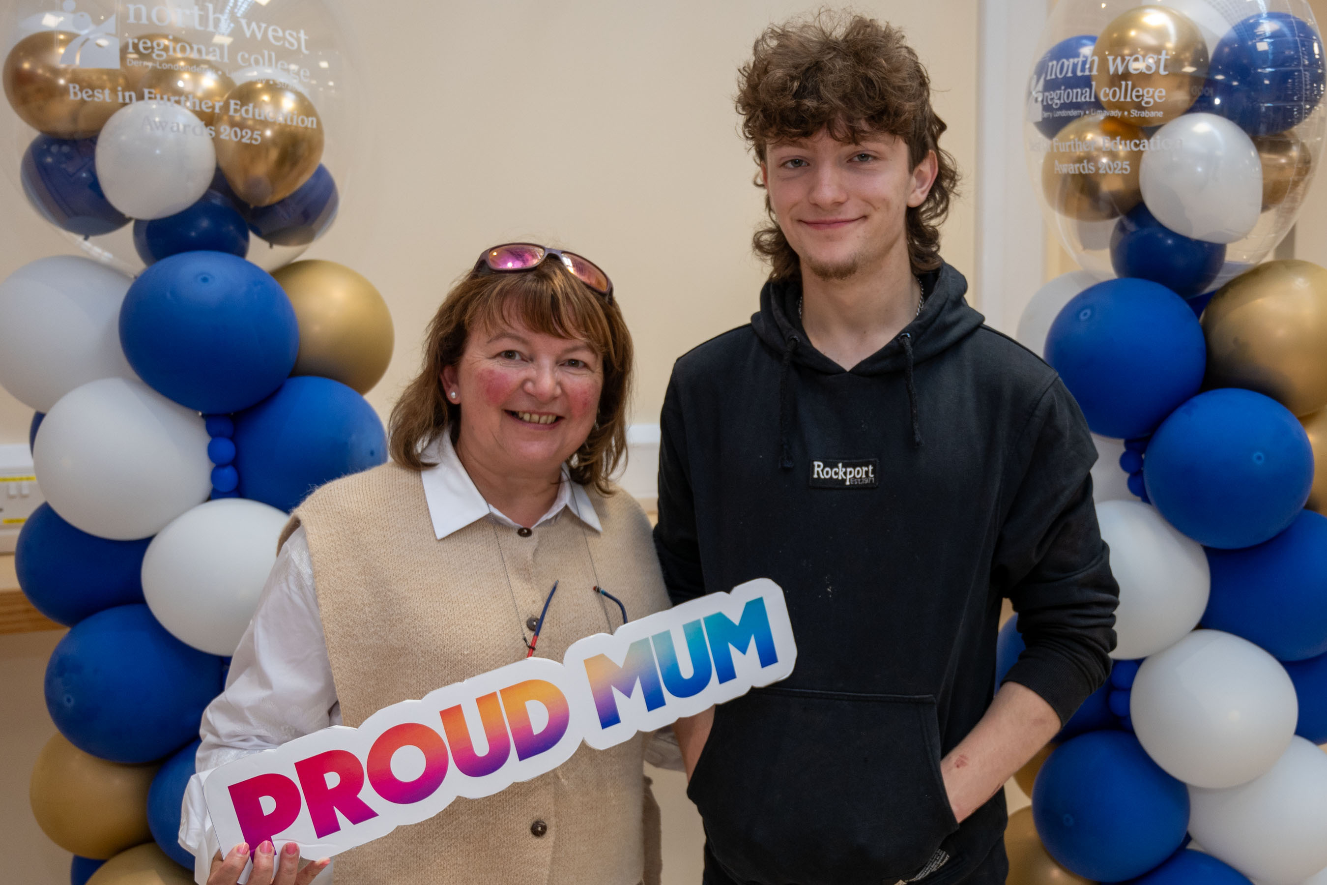 Two people at celebration event. One is holding a certificate, one has a proud mum sign.