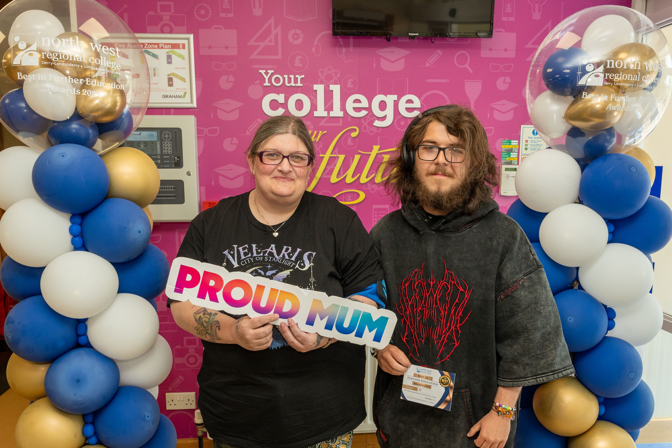 Two people at celebration event. One is holding a sign that says proud mum