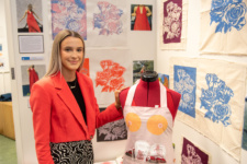 student pictured with artwork on the wall, beside a mannequin dressed in an apron that is part of the art