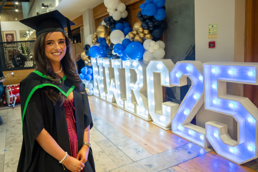 female graduate standing in front of large light-up lettering which reads NWRC25