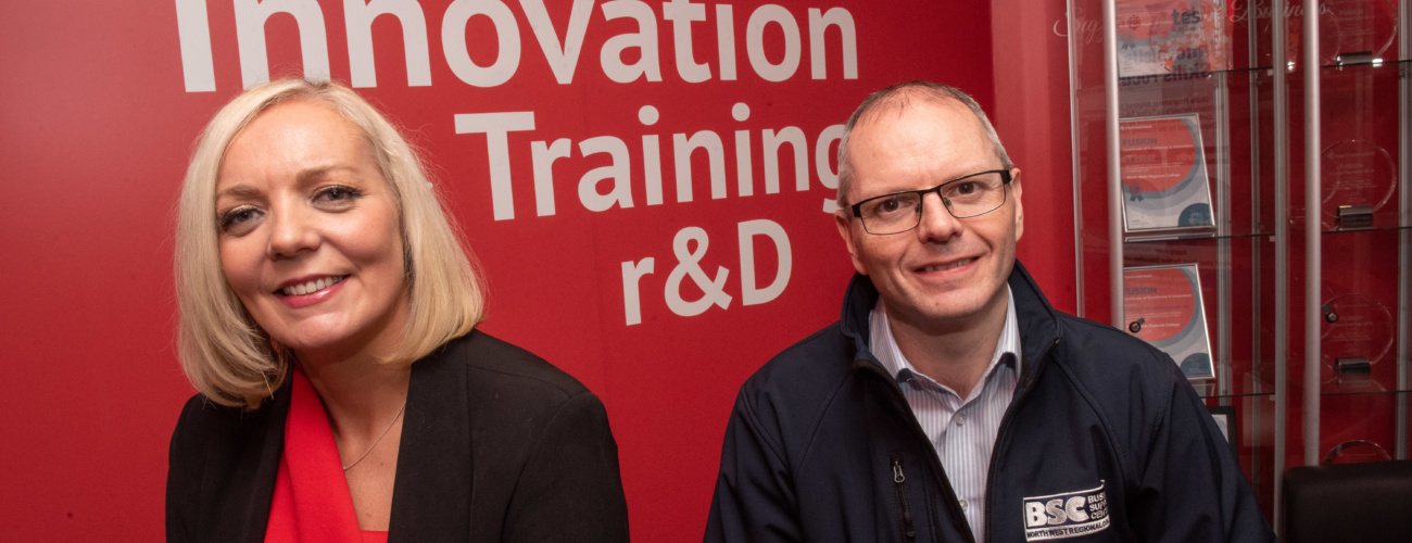 Man and woman smiling and sitting infront of a red wall with white writing on it