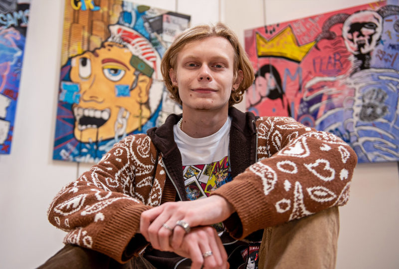 Art & Design student sits in front of his artwork displayed on the walls behind him