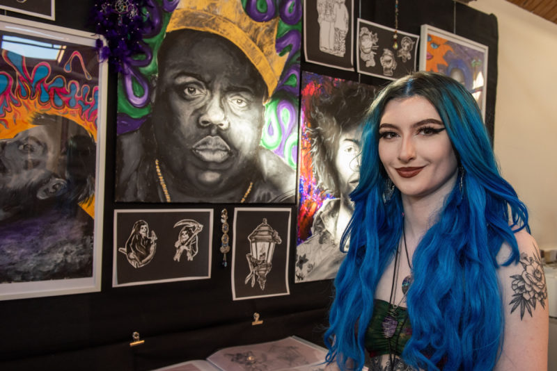 Female student with blue girl pictured in Art & Design studio in front of her painting displayed on the wall