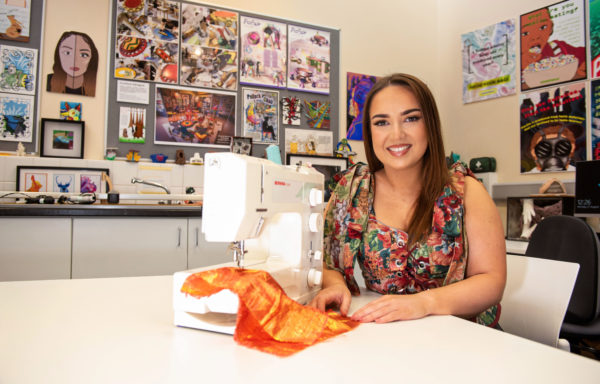 Young female smiling, sitting in Art classroom, using a sewing machine with paintings on the walls behind her