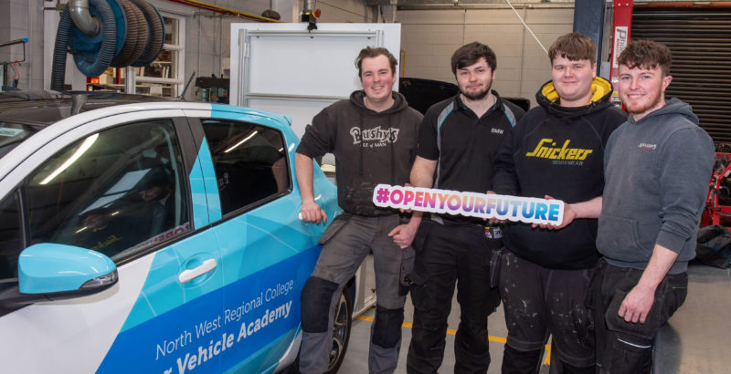 Four young men stand beside a car in the NWRC Motor Vehicle workshop, holding sign which read