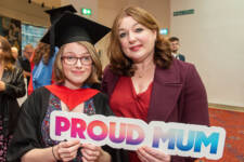 Young female graduate and her mother smile while holding Proud Mum cut out