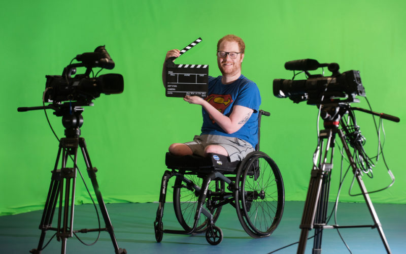 Male Media student in wheelchair holding movie clapper, in front of green screen with cameras
