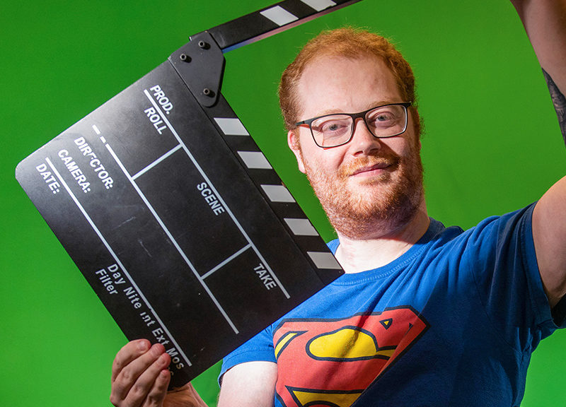 Ryan O'Connor, a male media student, sits in a wheelchair holding a movie clacker in front of a green screen