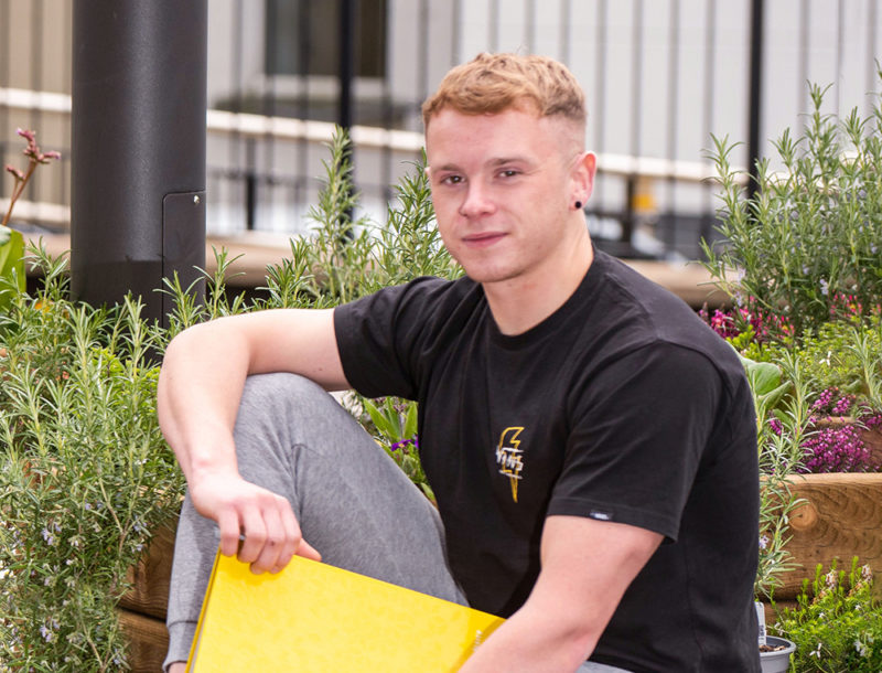 male student on campus sitting among flowers holding yellow lever-arch folder